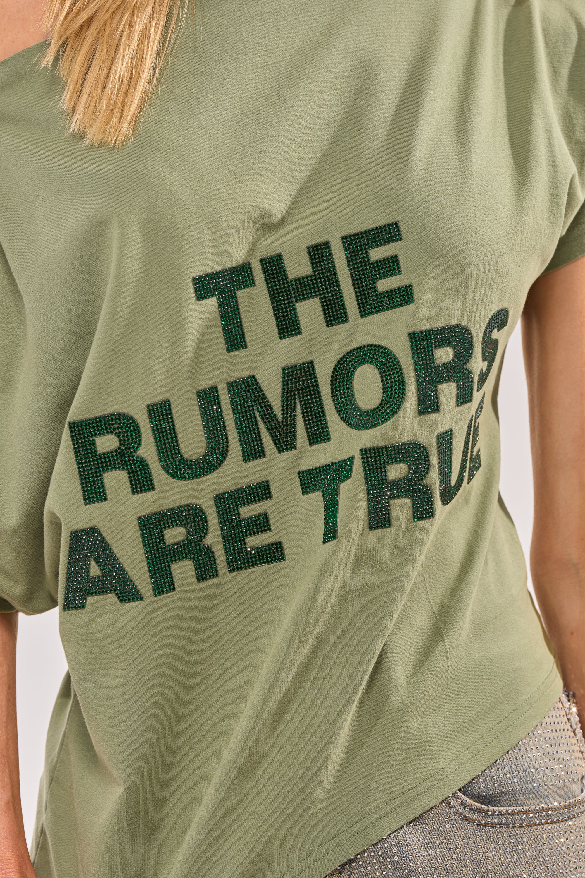 A person wearing the THE RUMORS ARE TRUE OFF THE SHOULDER OLIVE TEE, featuring bold dark green "THE RUMORS ARE TRUE" text. Only part of their upper body is shown. 6