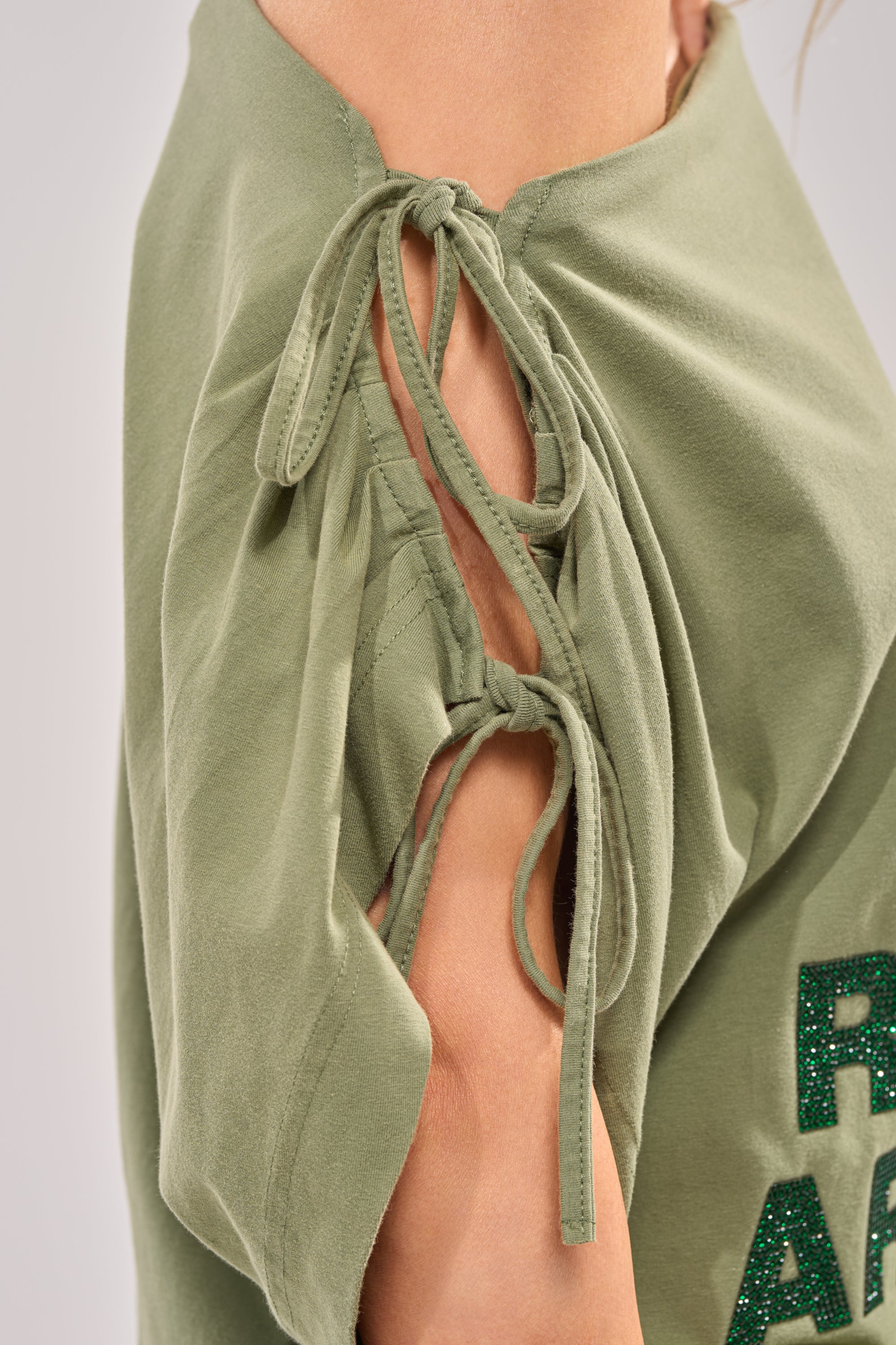 Close-up of THE RUMORS ARE TRUE OFF THE SHOULDER OLIVE TEE featuring green graphic rhinestones, two fabric ties on the seam revealing skin, and glittery text on the lower right for a bold streetwear vibe. 7