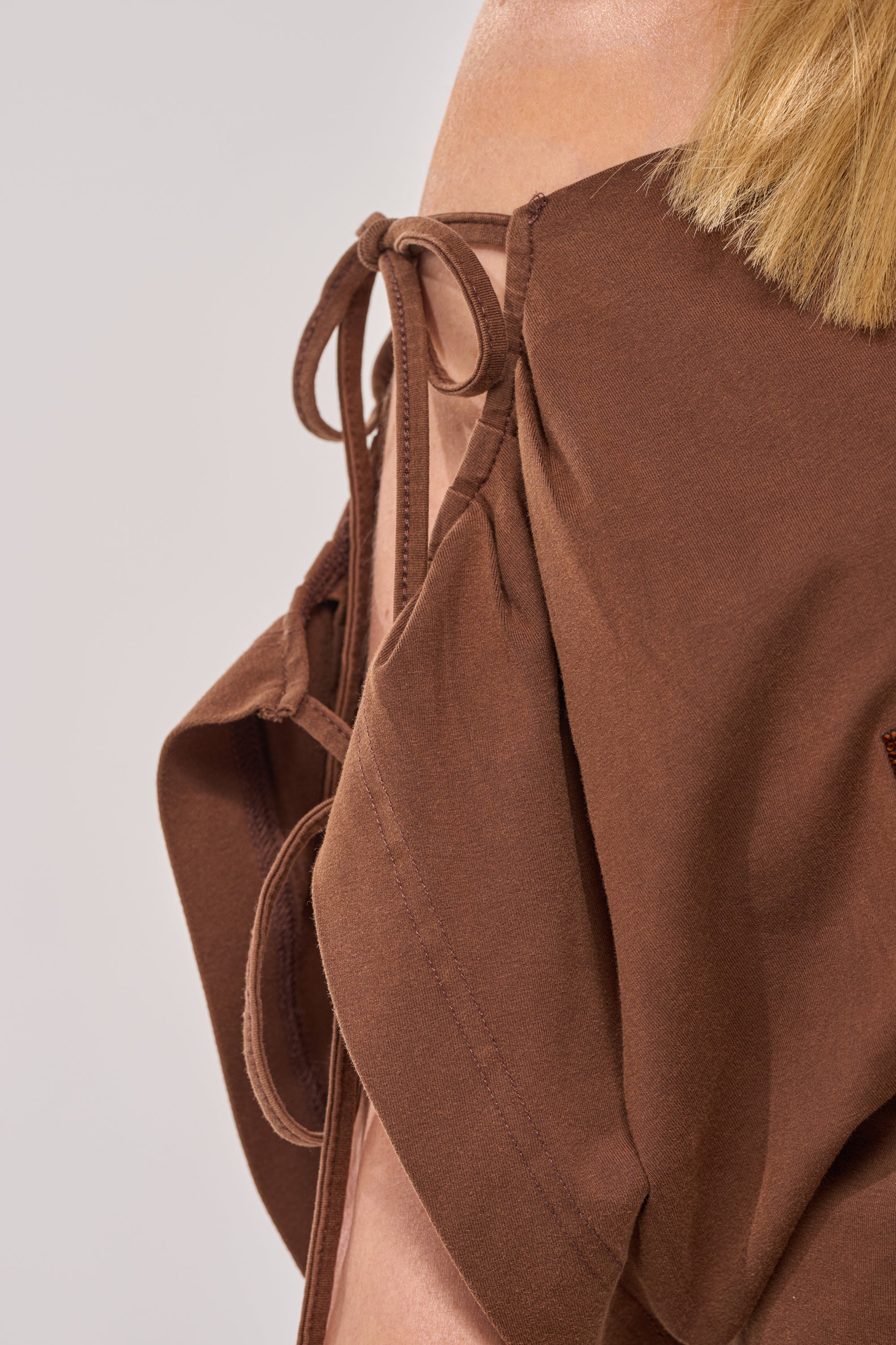 Close-up of a person wearing the FCK A DAY JOB OFF THE SHOULDER BROWN TEE, featuring tie-up straps and soft cotton fabric. The image highlights garment details and part of the person's blond hair. 10