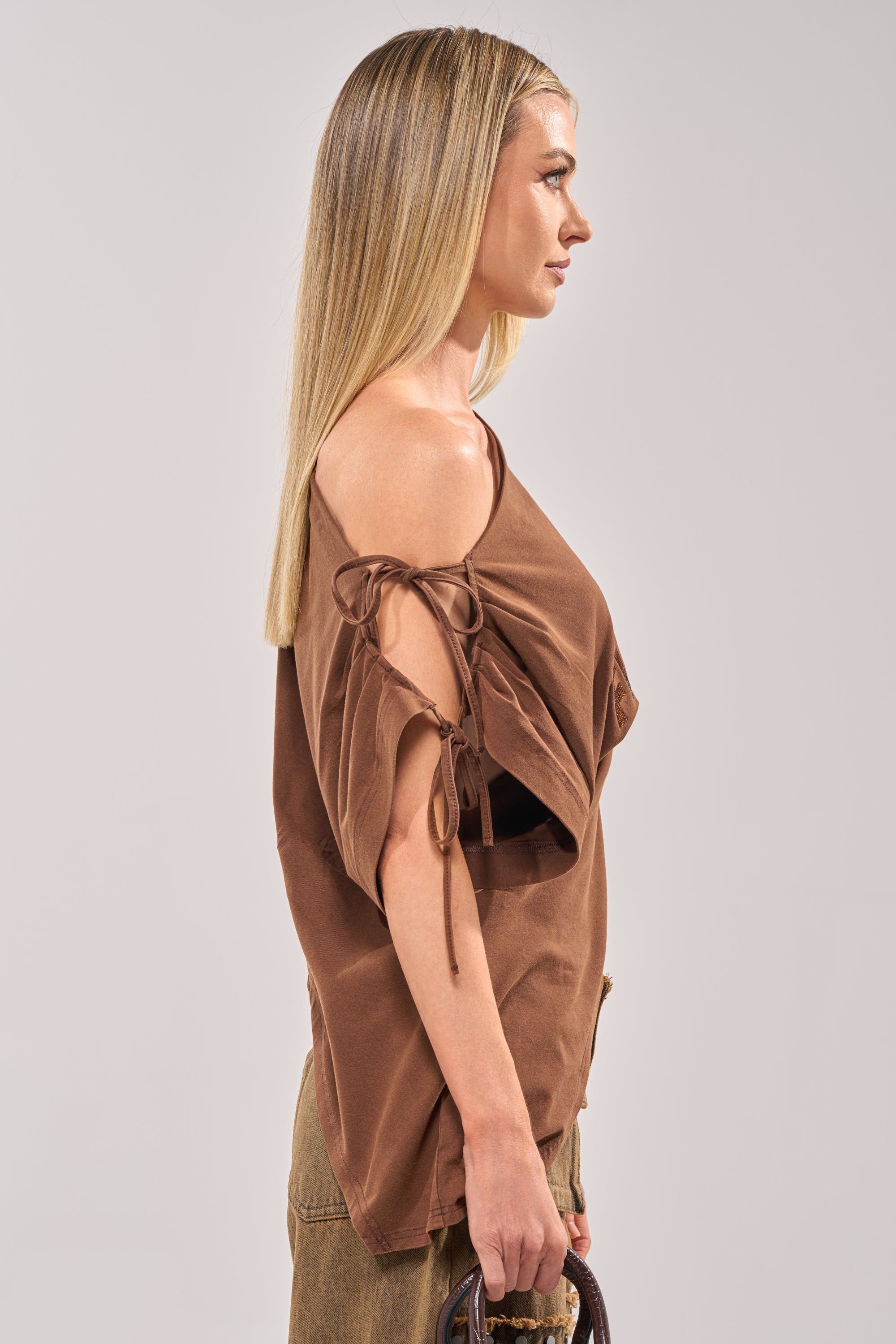 A woman with straight blonde hair stands in profile wearing the FCK A DAY JOB OFF THE SHOULDER BROWN TEE and khaki pants. She carries a brown handle bag and looks ahead against a plain light background. 5