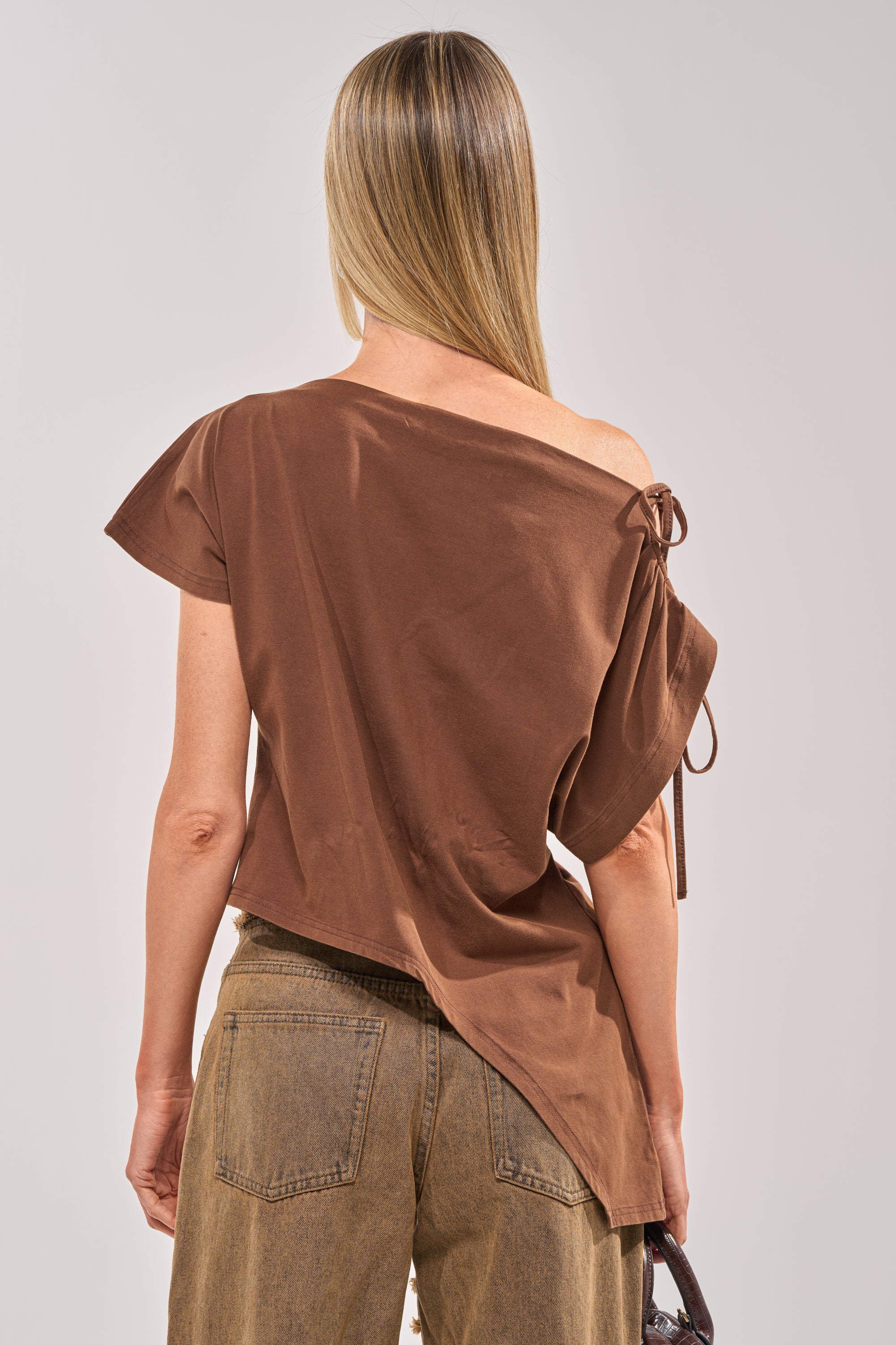 A woman with straight blonde hair wears the FCK A DAY JOB OFF THE SHOULDER BROWN TEE as an asymmetrical top, paired with high-waisted brown jeans. Seen from behind, she holds a small bag in her left hand. 6