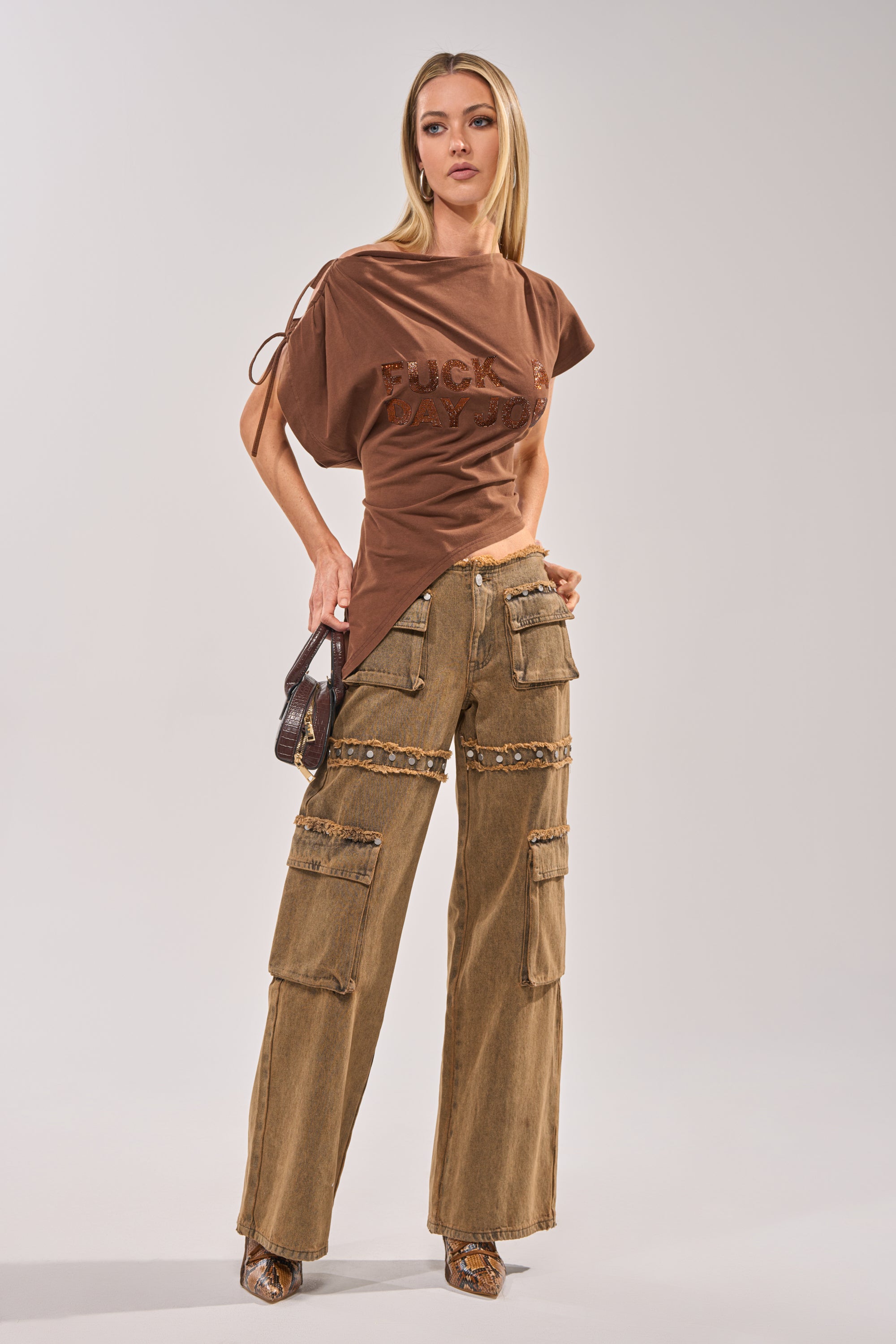 A woman poses against a plain background wearing the FCK A DAY JOB OFF THE SHOULDER BROWN TEE and tan cargo pants with snake-print details, accessorized with a small handbag and heeled shoes for an ideal festival streetwear look. 8
