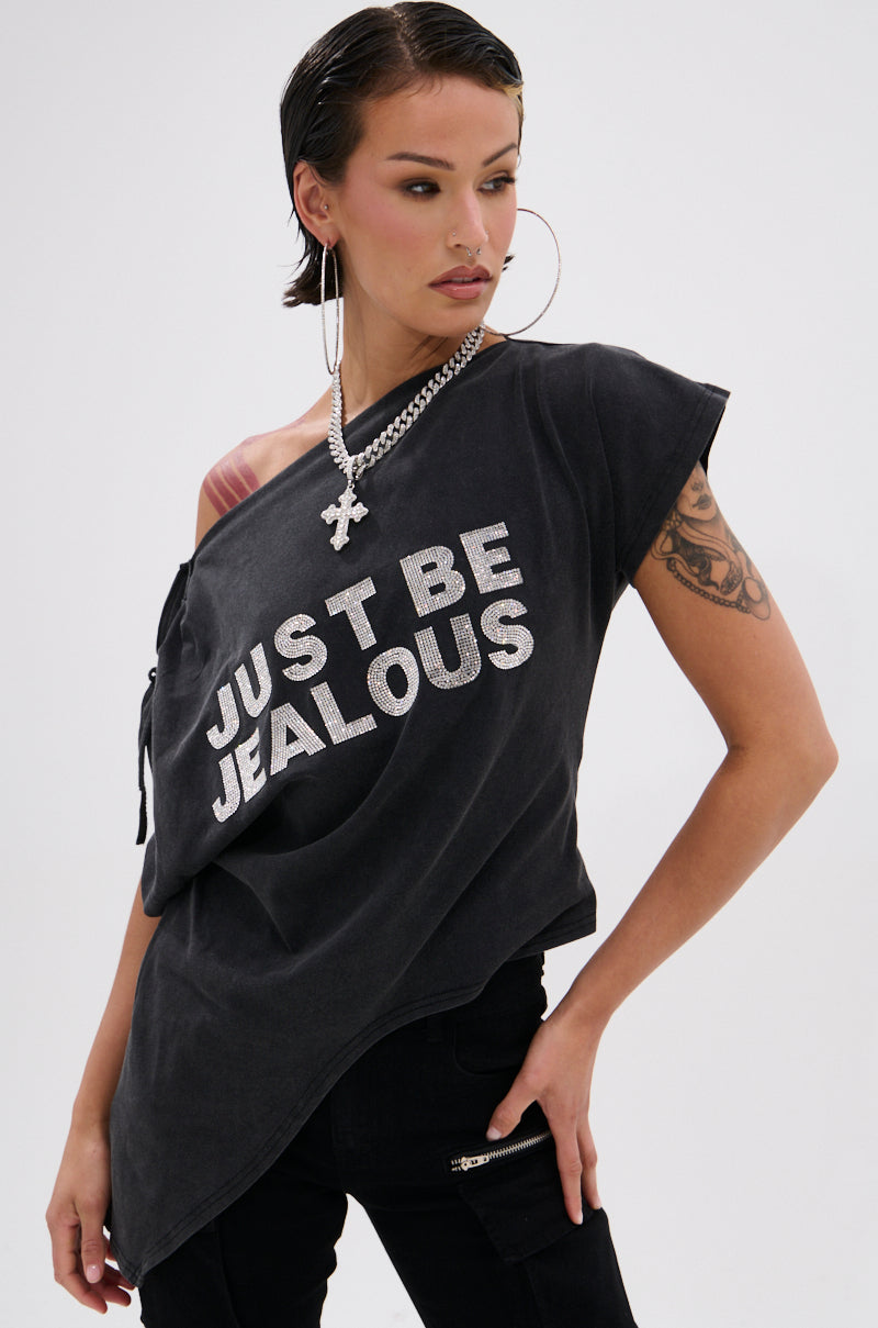 A person with short dark hair and large hoop earrings wears the JUST BE JEALOUS OFF THE SHOULDER TEE, a chunky chain necklace, and black pants, posing confidently with one hand on their hip.