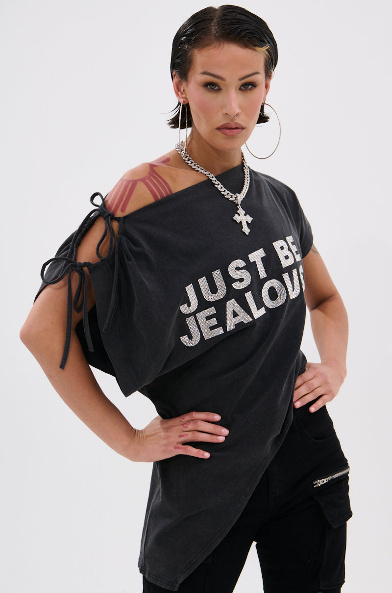 A confident woman with slicked-back hair and bold makeup models the JUST BE JEALOUS OFF THE SHOULDER TEE, featuring silver lettering. She pairs it with black pants, hoop earrings, a chain necklace, and stands with one hand on her hip.