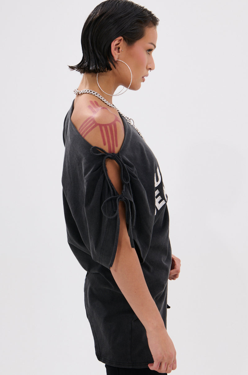 A person with short dark hair stands in profile, wearing a relaxed fit JUST BE JEALOUS OFF THE SHOULDER TEE with open sleeves tied by strings, showcasing a large geometric red shoulder tattoo, hoop earrings, and a chain necklace.