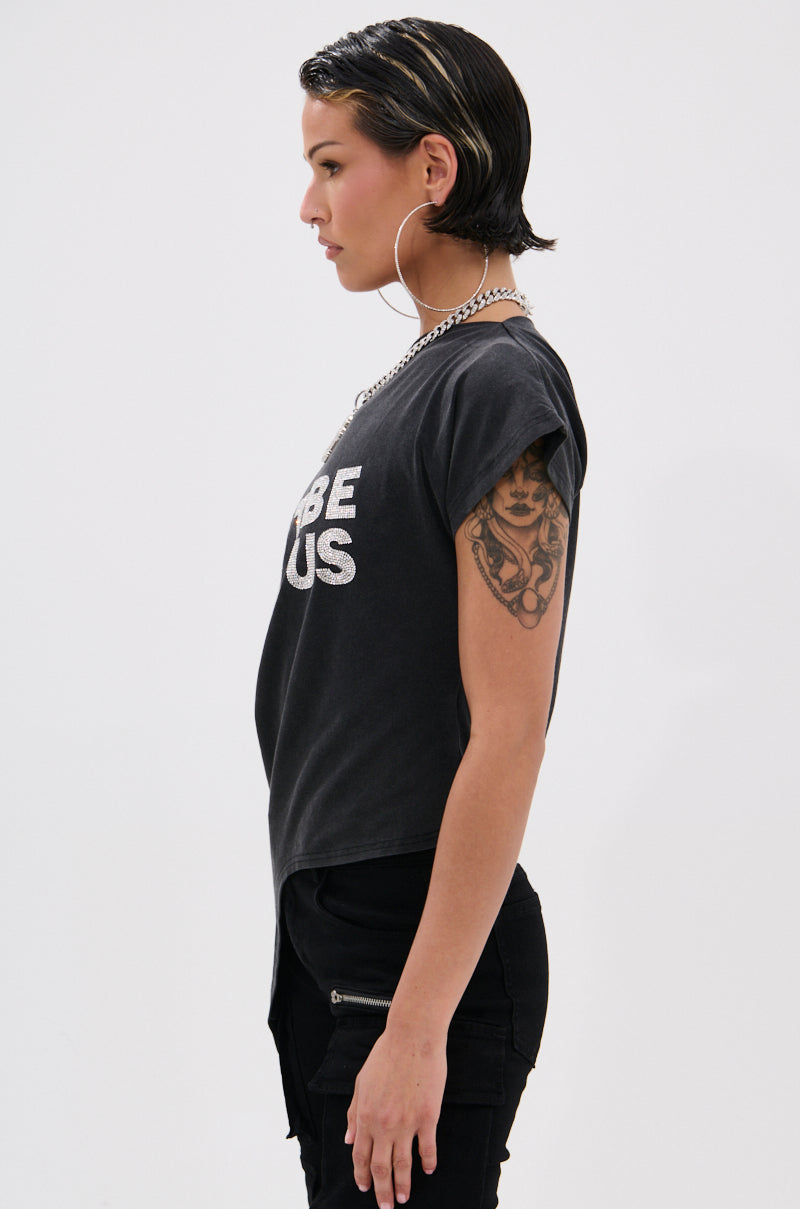 A person with short dark hair, large hoop earrings, necklace, and an off-the-shoulder black "JUST BE JEALOUS" tee stands in profile against a plain background. They have a visible upper arm tattoo.