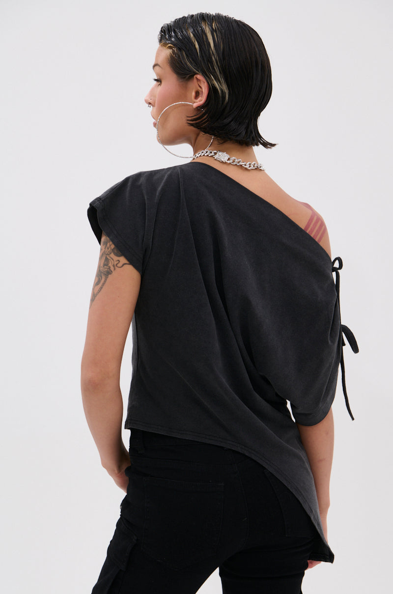 A person with short, wet-look hair wears the relaxed fit JUST BE JEALOUS OFF THE SHOULDER TEE tied at the shoulder, black pants, and a silver chain necklace. Slightly turned away, their pose highlights a tattooed arm.