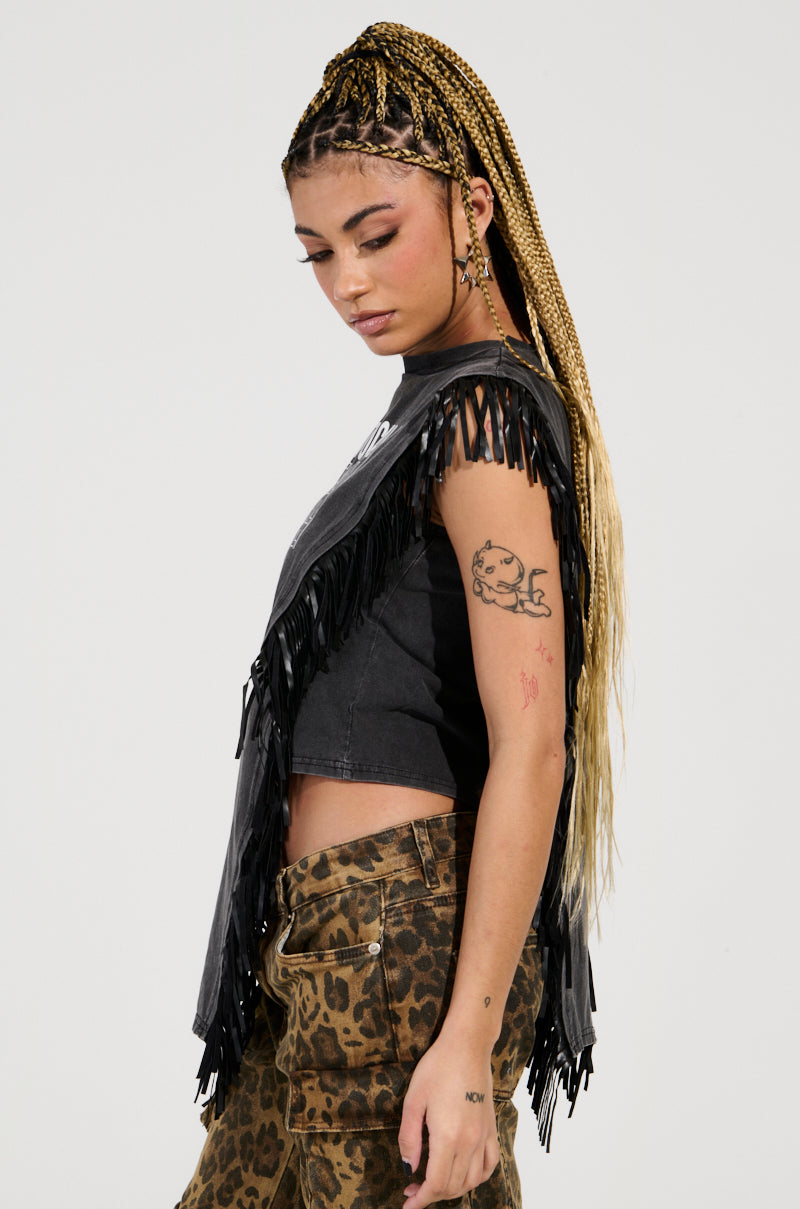A woman with long braided hair wears an ANTISOCIAL FRINGE DETAIL TSHIRT and leopard-print pants. With tattoos on her arm, she gazes downward against a plain background.