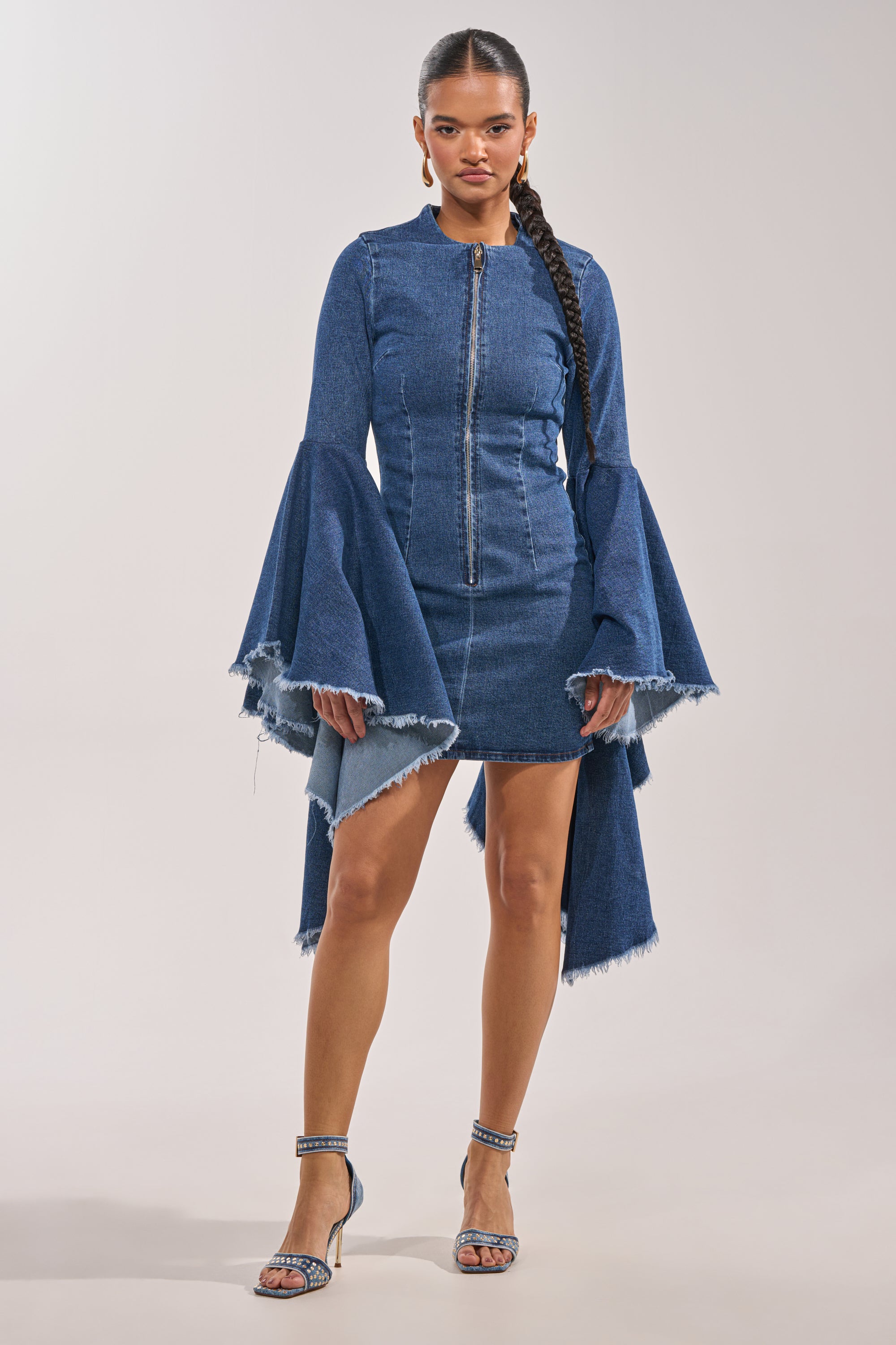 A woman in the WALK IN THE PARK DENIM MINI DRESS, featuring tiered bell sleeves and a frayed hem, pairs it with blue high-heeled sandals. She wears her hair in a long braid and exudes confidence. 1