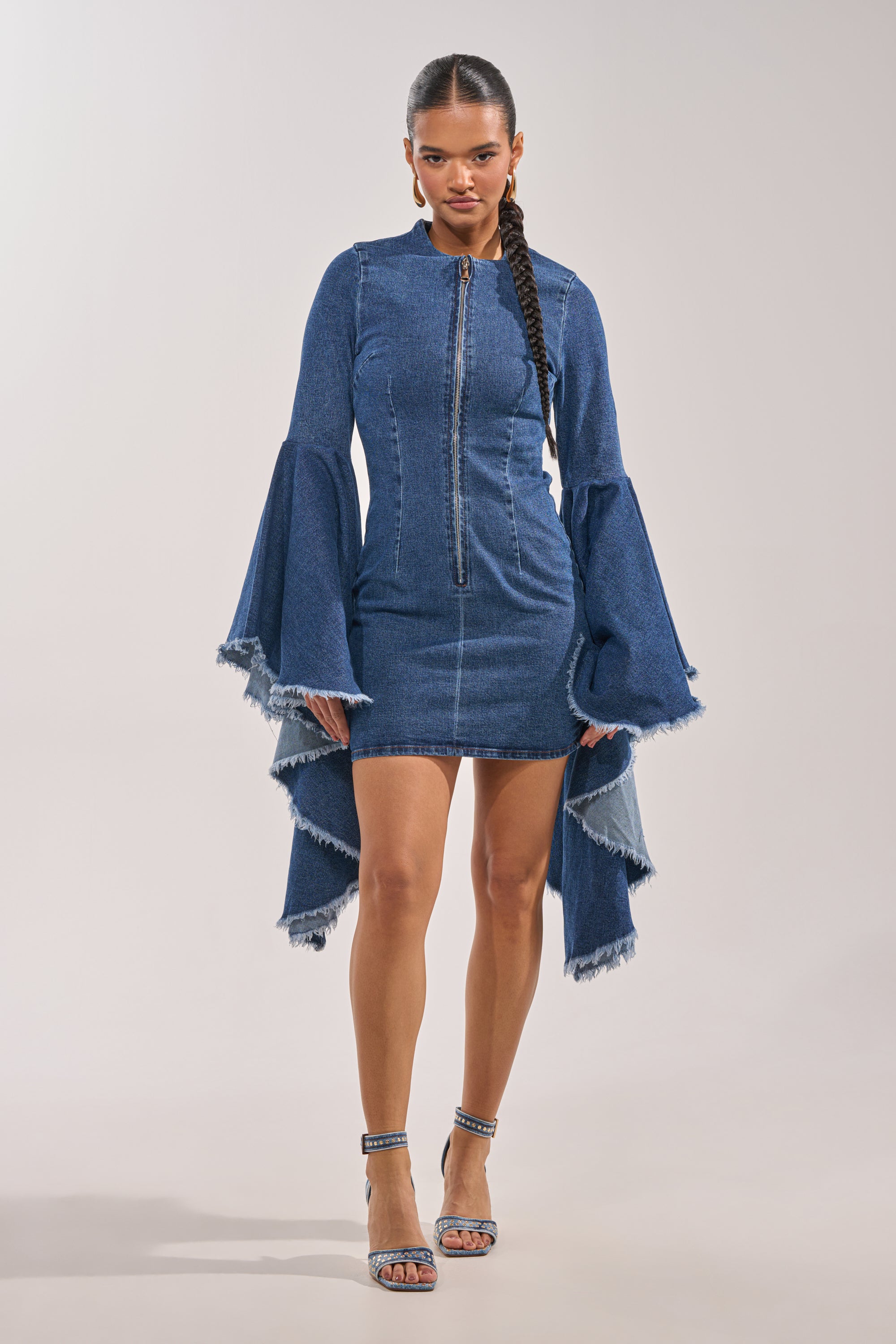A woman faces forward in the WALK IN THE PARK DENIM MINI DRESS, featuring tiered bell sleeves and a frayed hemline. She styles it with a long braid and coordinating denim high-heeled sandals. 2