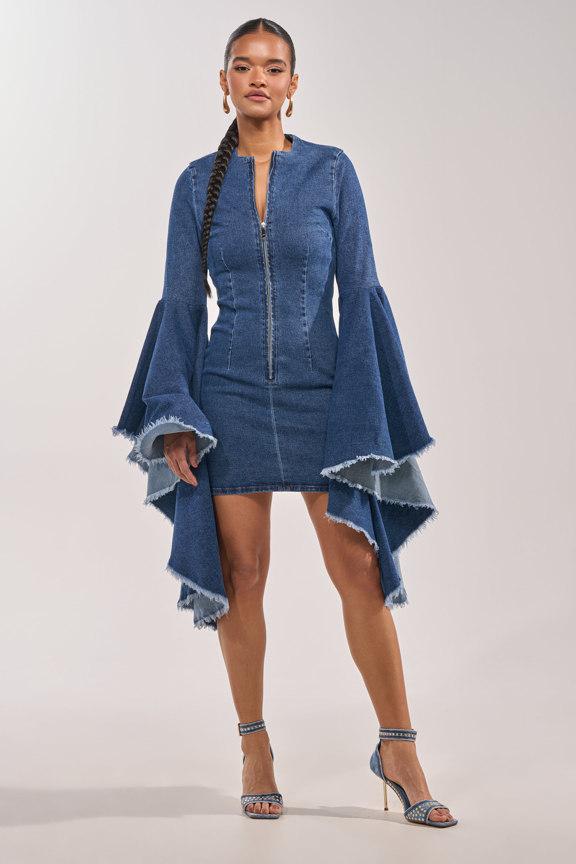 A woman stands confidently in the WALK IN THE PARK DENIM MINI DRESS, featuring dramatic bell sleeves, an exposed front zip, and frayed edges, paired with blue high-heeled sandals against a neutral background. 3