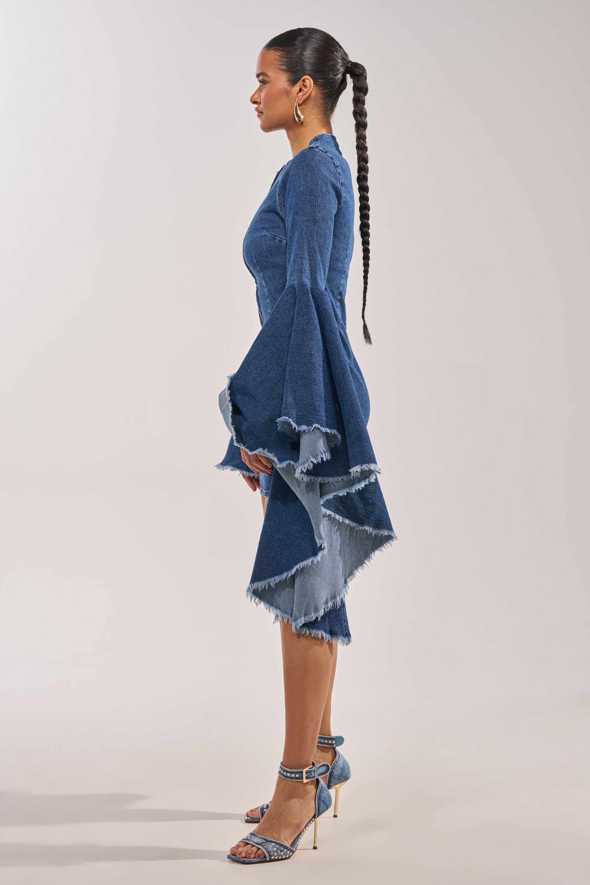 A woman stands in profile wearing the WALK IN THE PARK DENIM MINI DRESS, featuring tiered bell sleeves and a frayed hemline. She styles it with a long braided ponytail and high-heeled denim-style sandals. 4