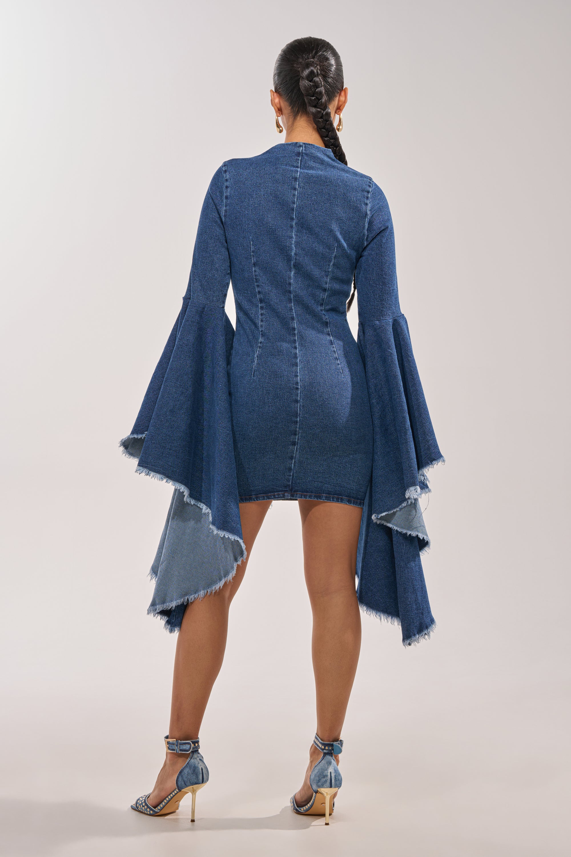 A woman with braided hair stands with her back to the camera, wearing the WALK IN THE PARK DENIM MINI DRESS featuring dramatic bell sleeves and frayed edges, paired with blue high-heeled sandals. 5