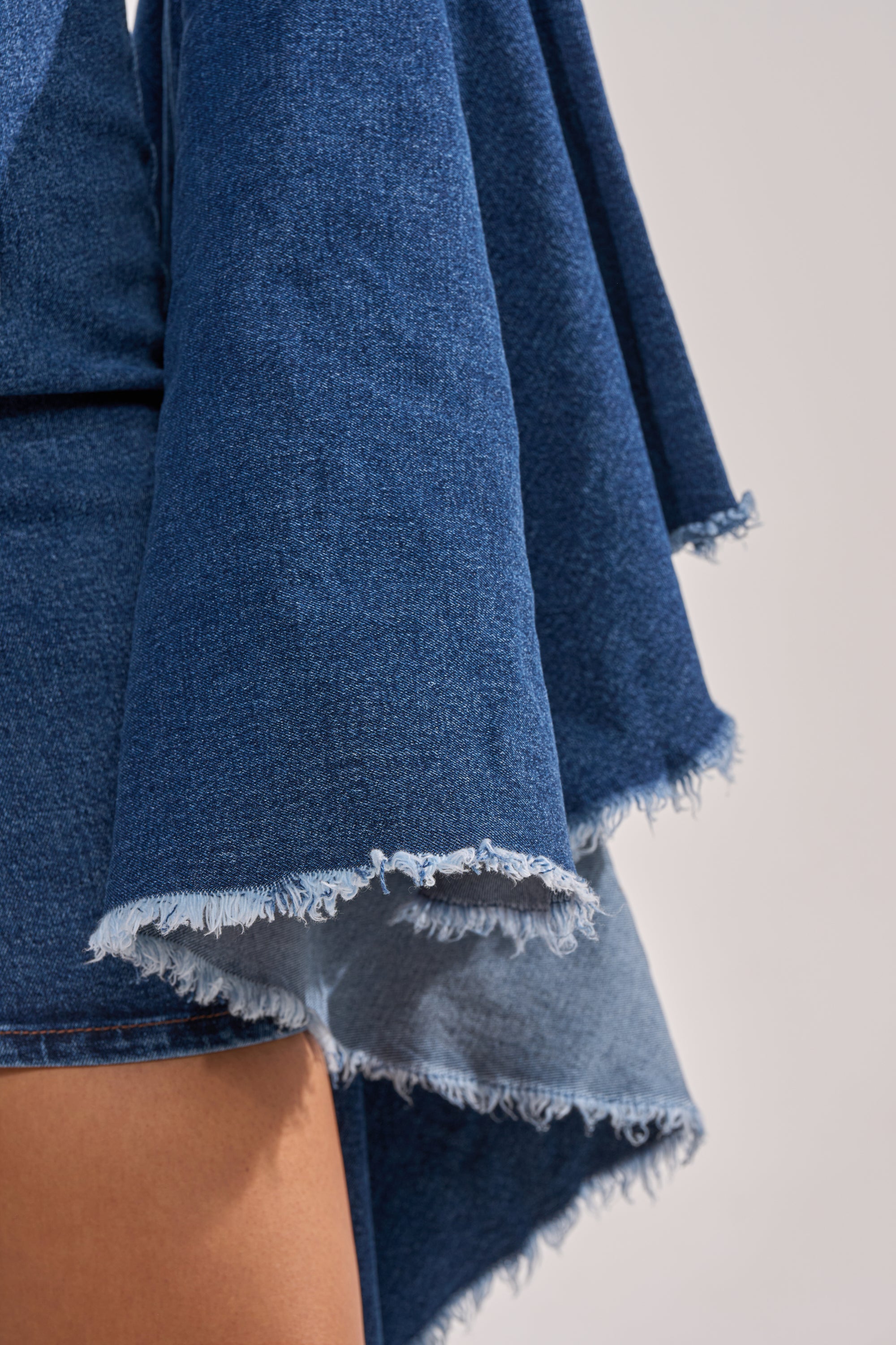 Close-up of a person wearing the WALK IN THE PARK DENIM MINI DRESS, featuring wide, frayed bell sleeves, textured blue denim, and an exposed front zip. Part of the wearer’s leg is visible in the lower left corner. 6