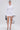 A woman faces forward in the TRACY DRAMATIC SLEEVE MINI DRESS, a fitted white stretch knit with bold black-trimmed bell sleeves. She carries a small black handbag, wears strappy high heels, and her hair is styled sleekly back.