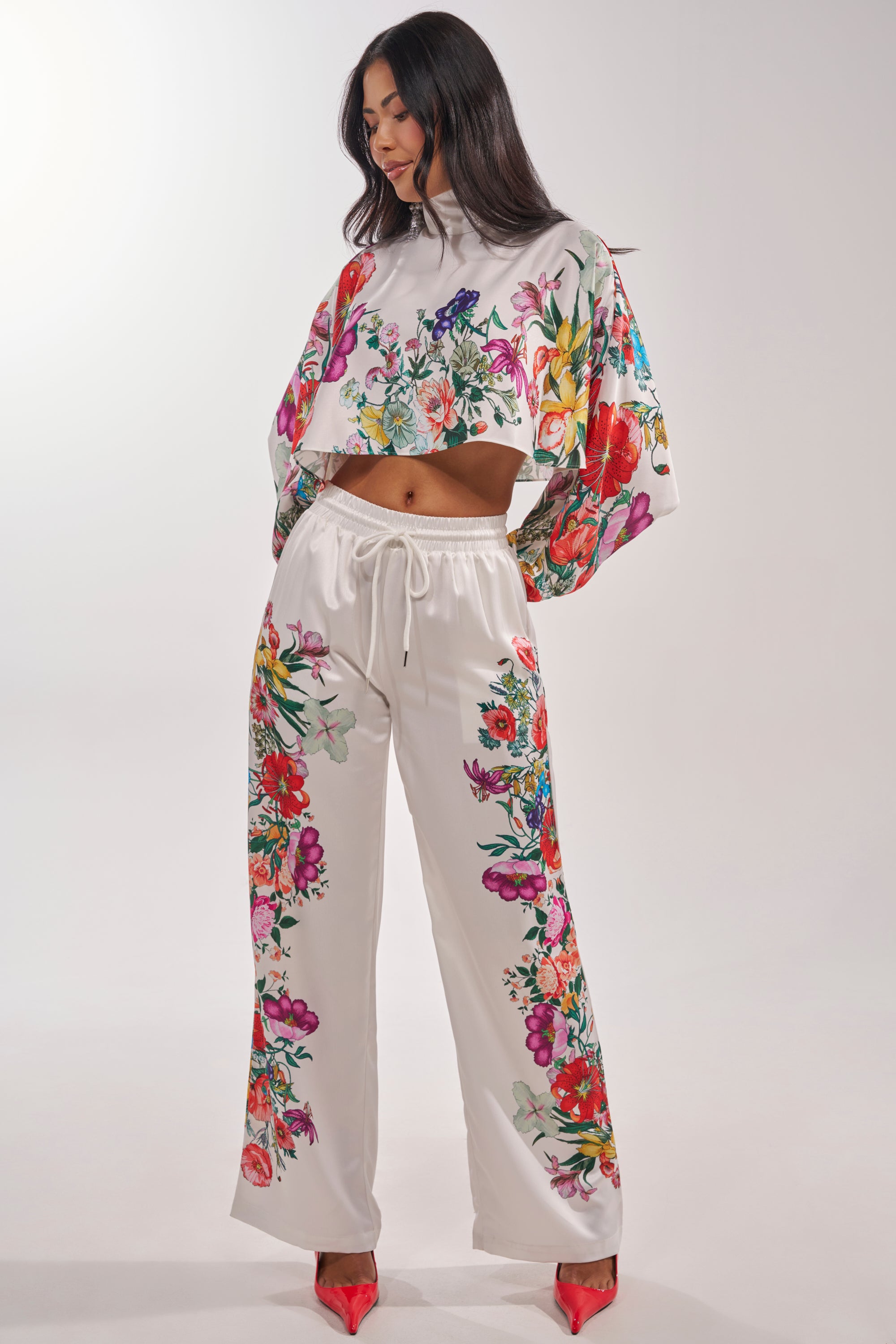 A woman models the FULL BLOOM STRAIGHT LEG TROUSERS in a floral print, paired with a white, long-sleeve crop top and red pointed heels, creating a fresh spring look against a light background. 2