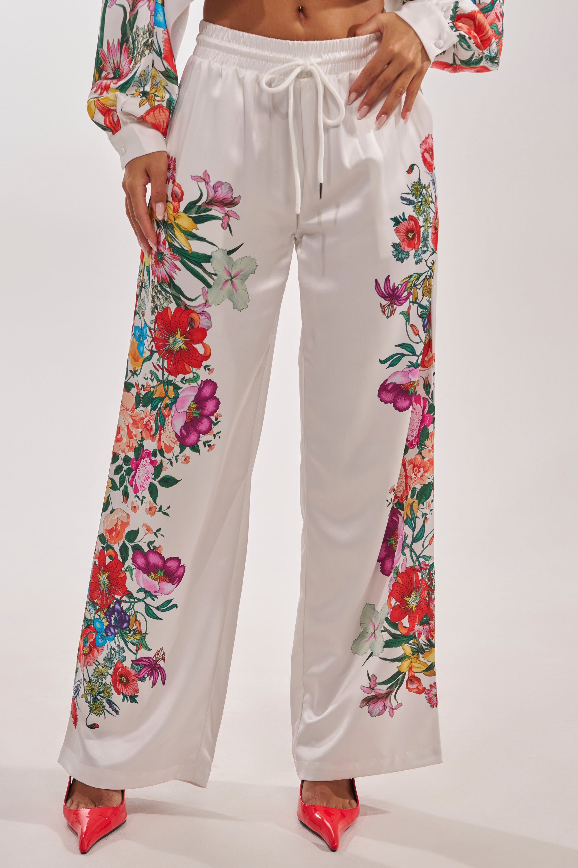 A person wears the FULL BLOOM STRAIGHT LEG TROUSERS with colorful floral prints and a drawstring waist, styled with matching long sleeves and bright red pointed shoes for a chic spring brunch look. 4