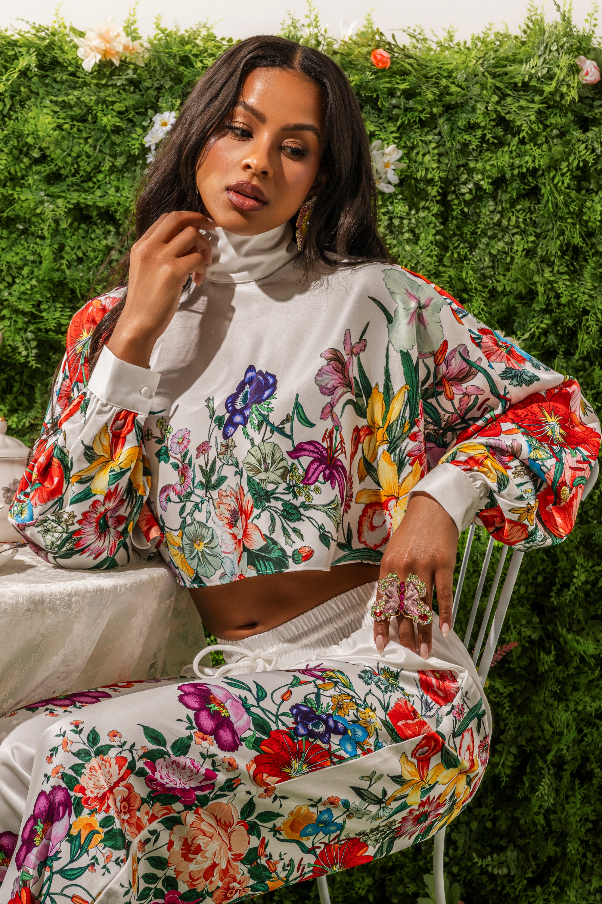A woman sits by a table, framed by green leaves and flowers, wearing the FULL BLOOM SATIN MOCK NECK BLOUSE IN WHITE with vibrant, colorful print pants.