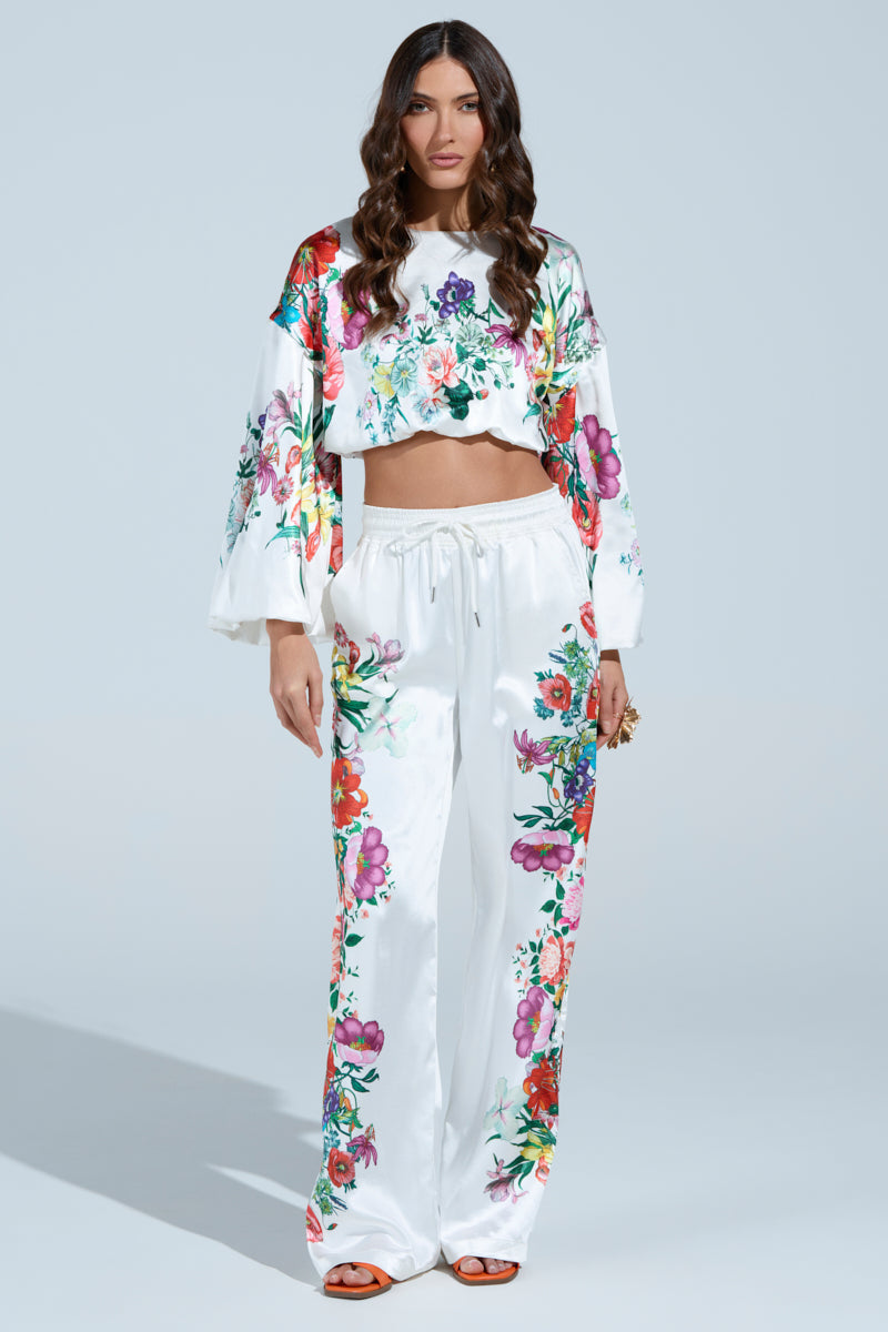 A woman with long wavy hair wears the FULL BLOOM STRAIGHT LEG TROUSERS and a vibrant floral crop top with bell sleeves. Posed confidently against a light background, she completes her look with rings and orange sandals.