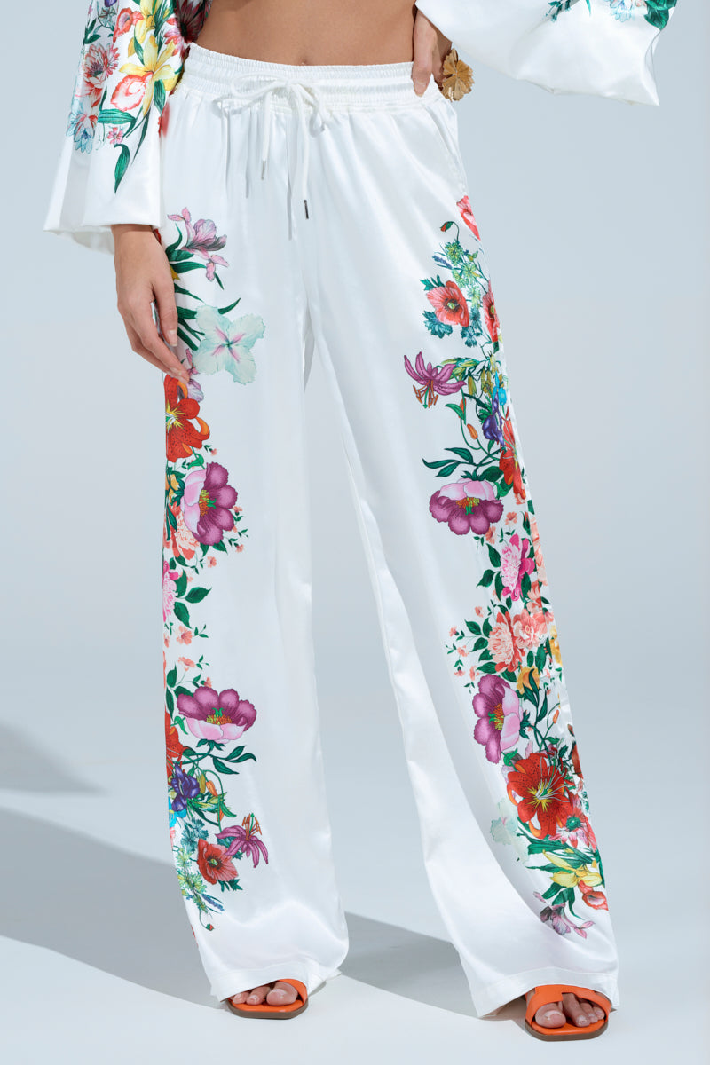 Dressed in the FULL BLOOM STRAIGHT LEG TROUSERS featuring a high-rise waist and colorful floral prints of red, pink, and purple, paired with a matching top with outstretched sleeves and orange sandals. They pose elegantly against a light blue background.