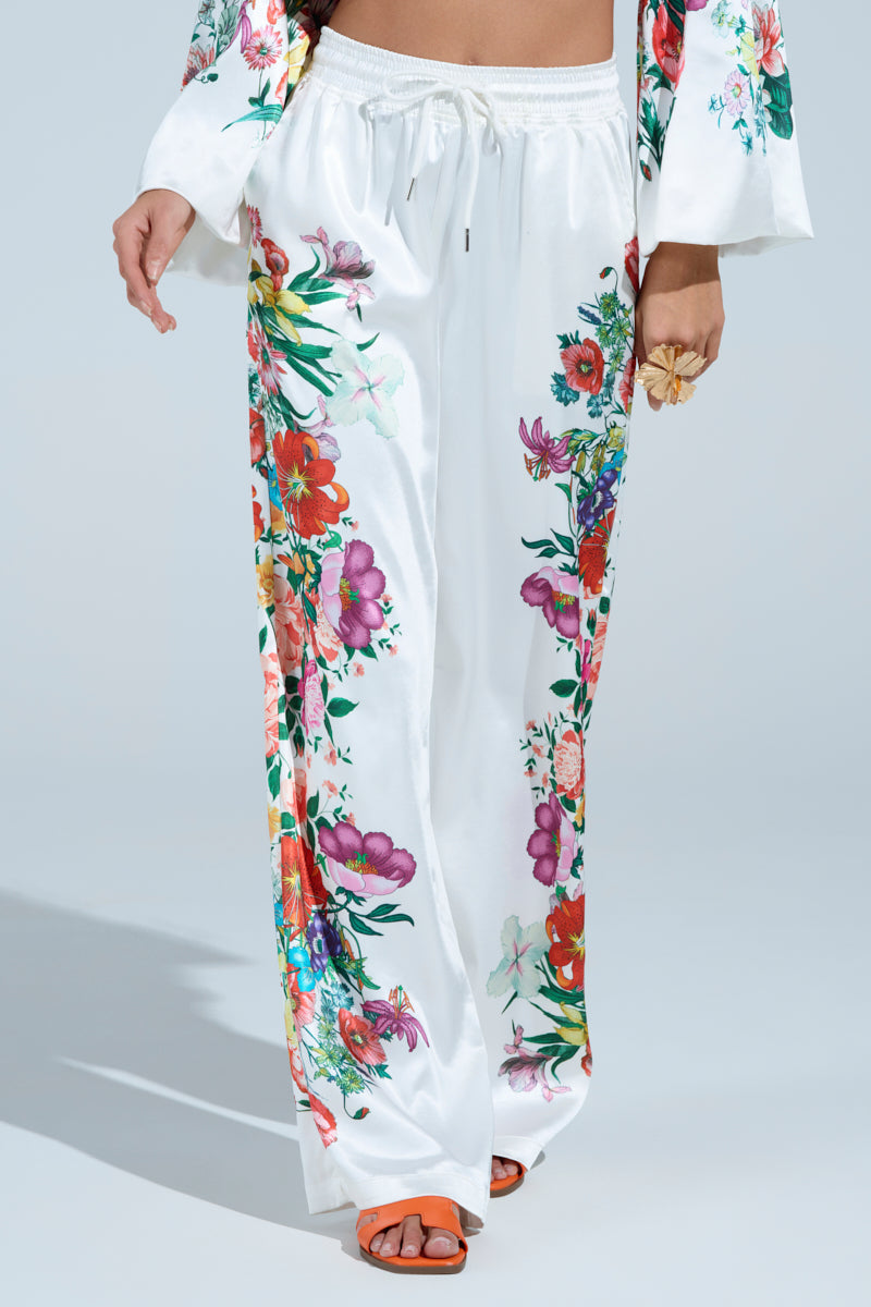 A person dressed in FULL BLOOM STRAIGHT LEG TROUSERS, featuring a high-rise waist, colorful floral print, elastic waistband, and drawstring, paired with a matching top and orange sandals. They hold a flower accessory in one hand.