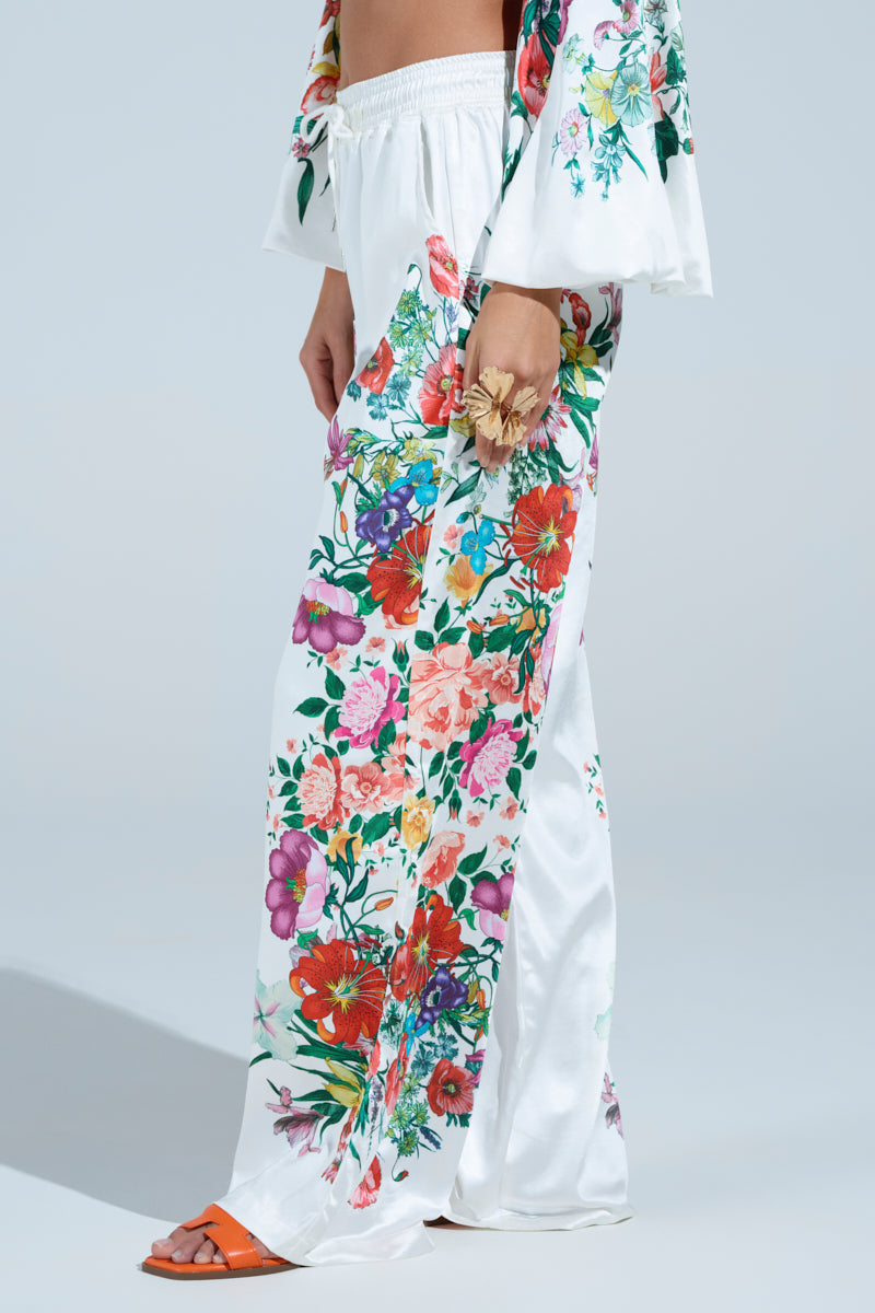 A person wearing FULL BLOOM STRAIGHT LEG TROUSERS, featuring a floral print and high-rise waist, holds a flower. Their outfit is beautifully completed with orange heeled sandals against a light, solid background that highlights the vibrant ensemble.