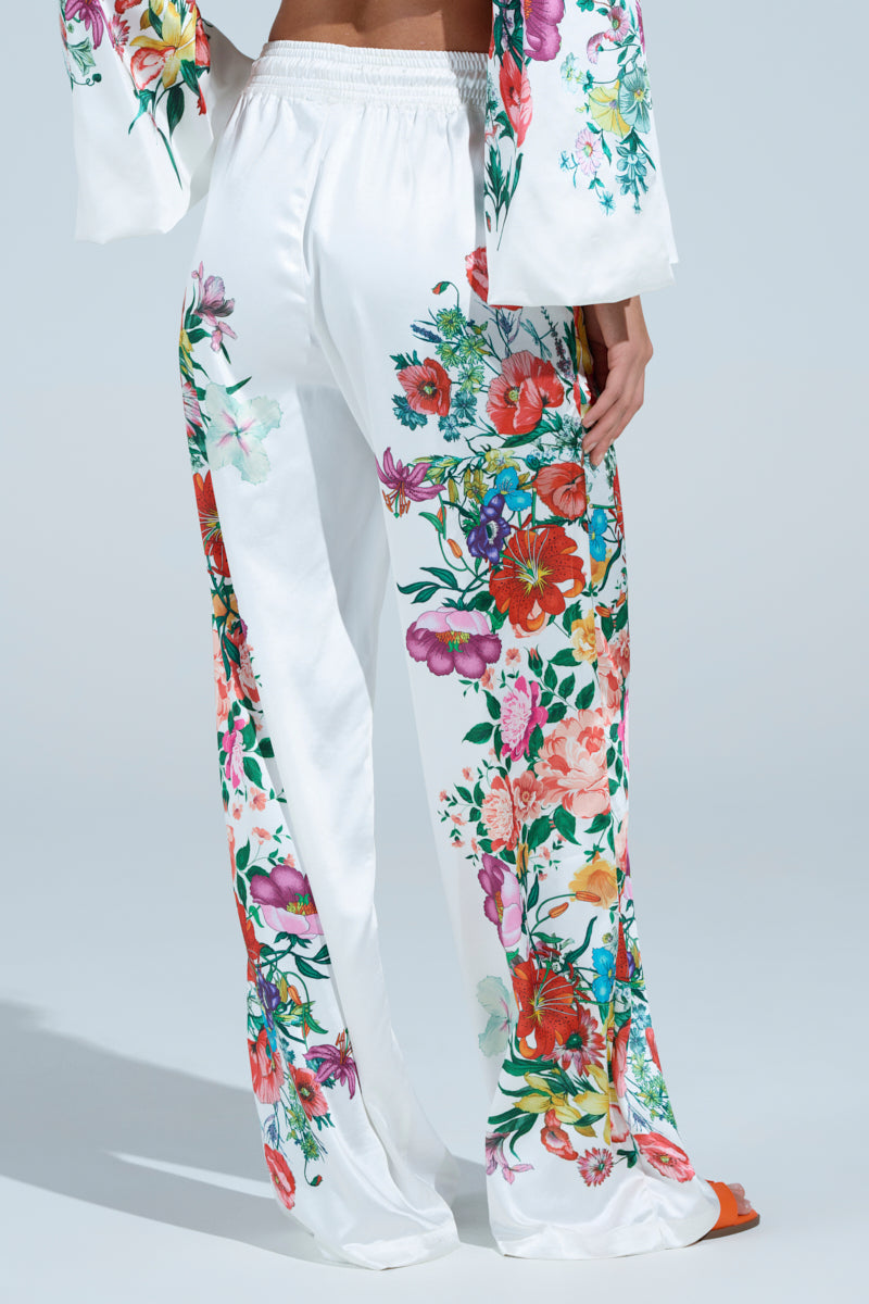 Wearing FULL BLOOM STRAIGHT LEG TROUSERS, a person showcases a vibrant floral print of red, pink, and orange blooms on white satin pants. The high-rise waistband adds elegance against the plain background.