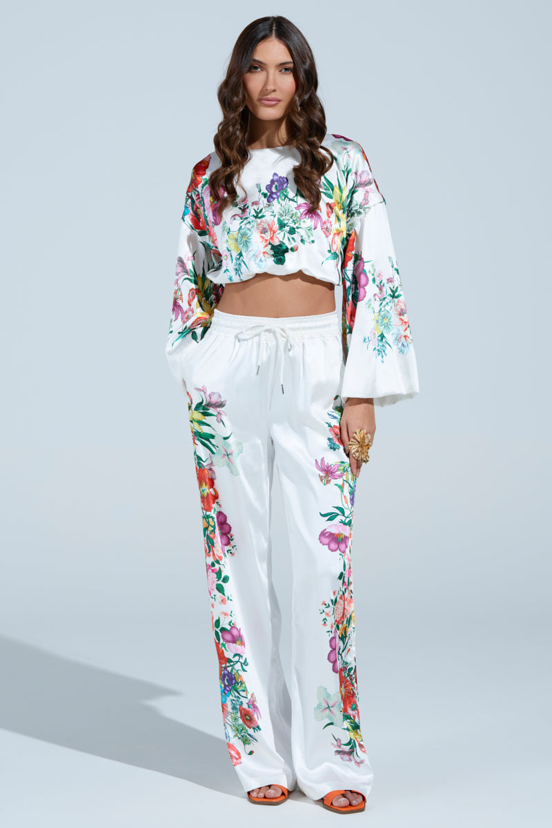 A woman in a floral-patterned loose outfit featuring a cropped top and satin FULL BLOOM STRAIGHT LEG TROUSERS poses confidently with hands in pockets against a minimalist light background. Her long, wavy hair adds elegance to the scene.