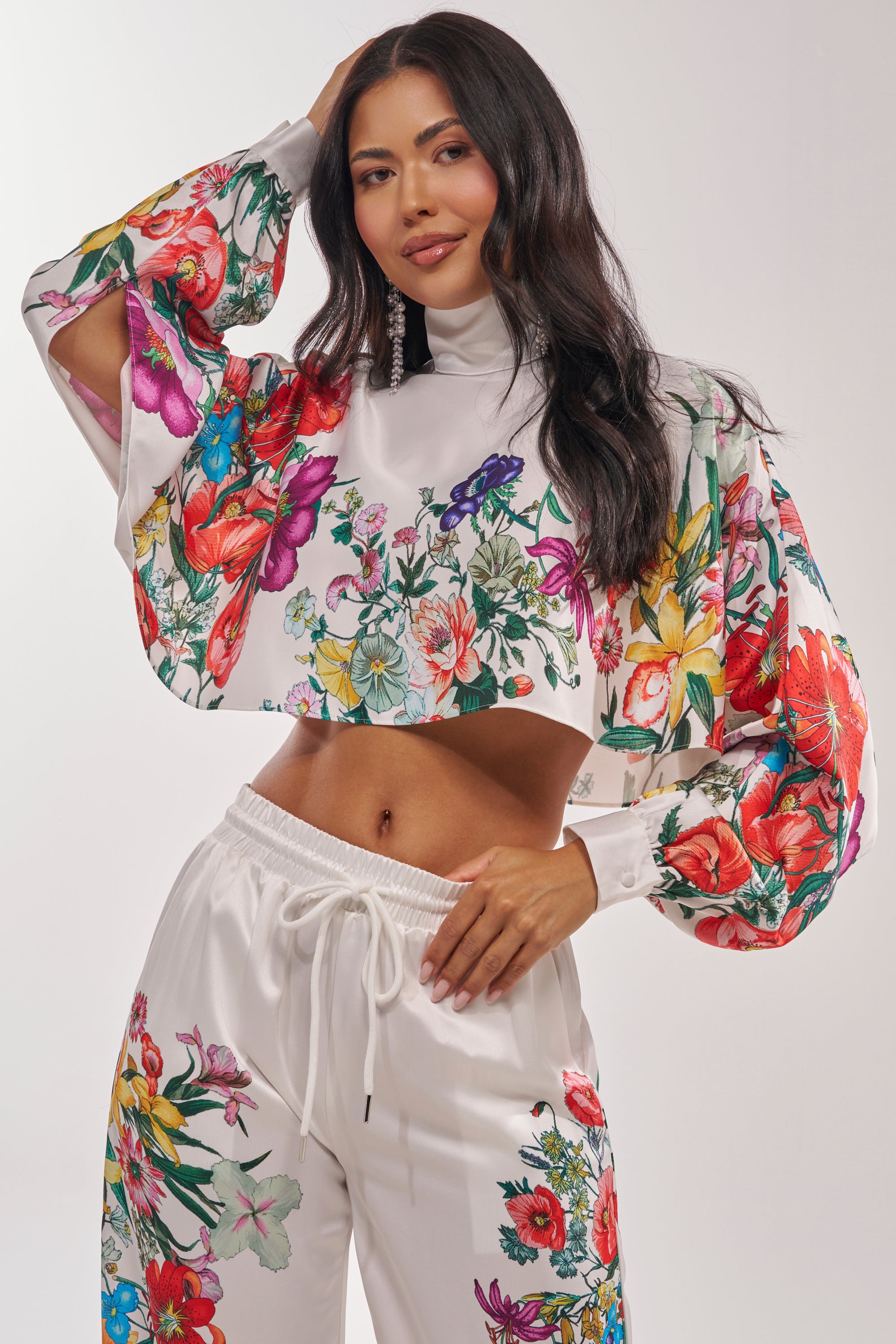A woman with long dark hair wears the FULL BLOOM SATIN MOCK NECK BLOUSE IN WHITE with a colorful floral print. She smiles confidently, posing with one hand on her hip and the other touching her head.