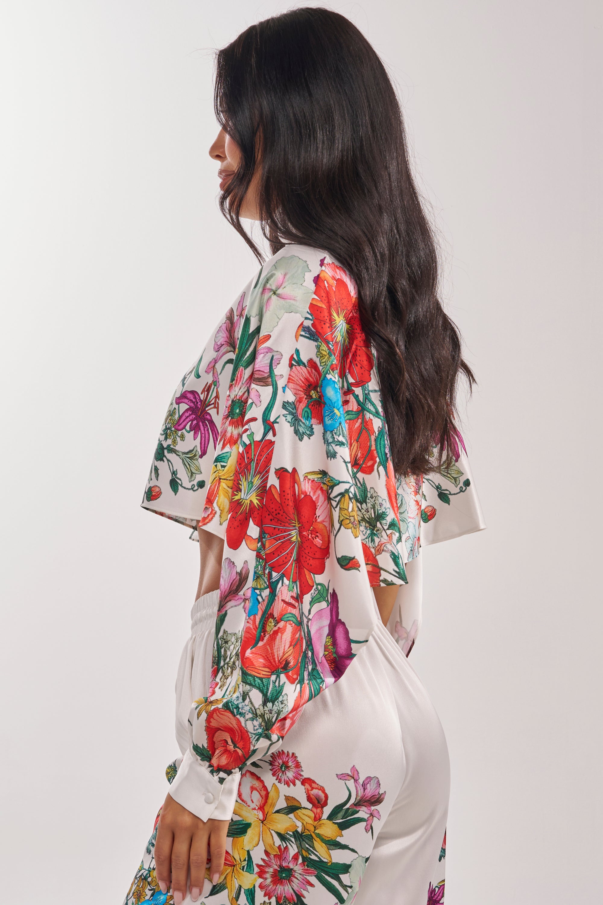 Woman with long, wavy dark hair wears the FULL BLOOM SATIN MOCK NECK BLOUSE IN WHITE, standing sideways against a plain light background.