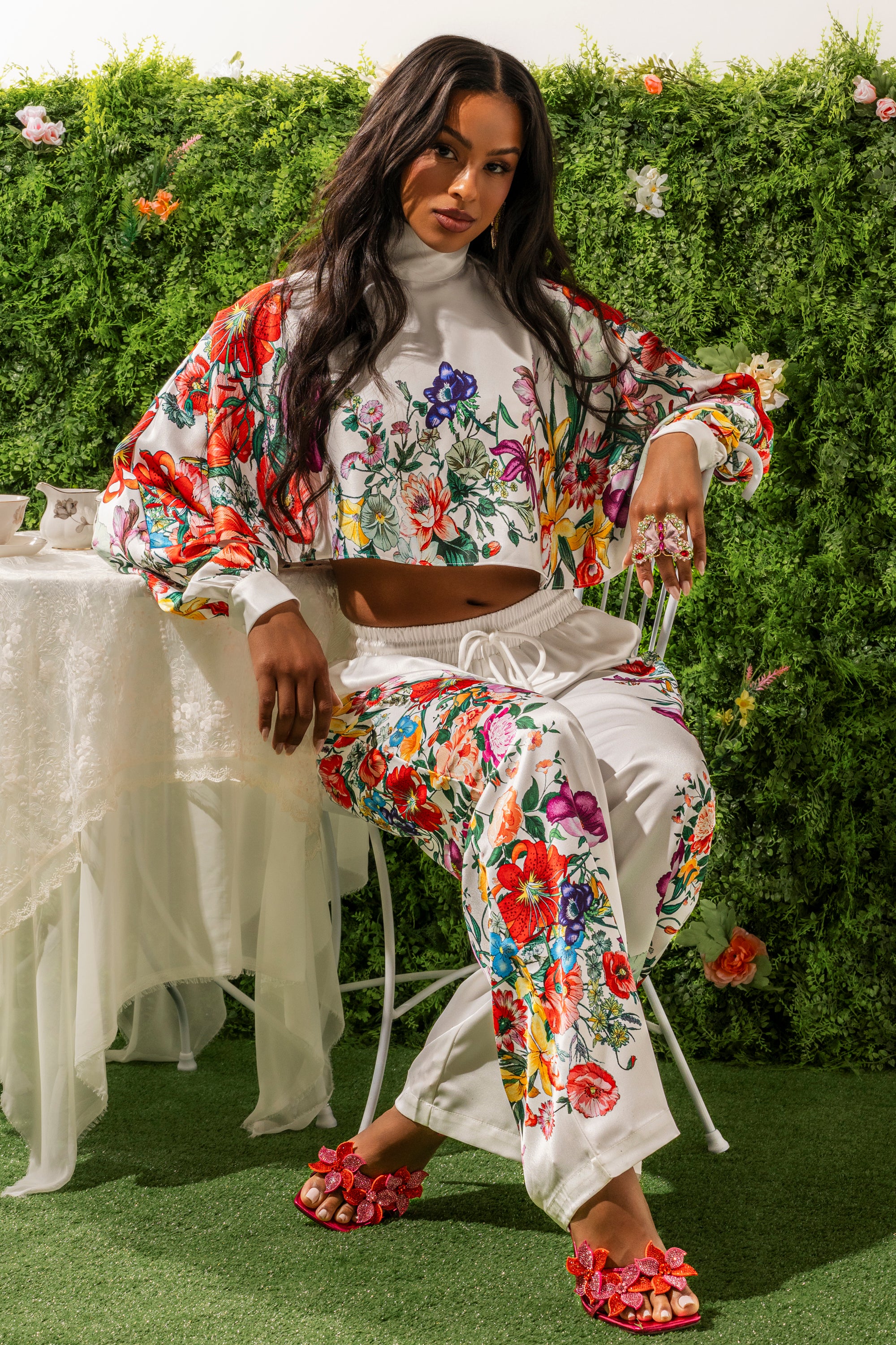 A woman sits on a white chair in a garden, wearing FULL BLOOM STRAIGHT LEG TROUSERS and a matching floral top—perfect for spring brunch. She has long dark hair, pink floral sandals, lush plants around her, and a lace-draped table nearby. 0