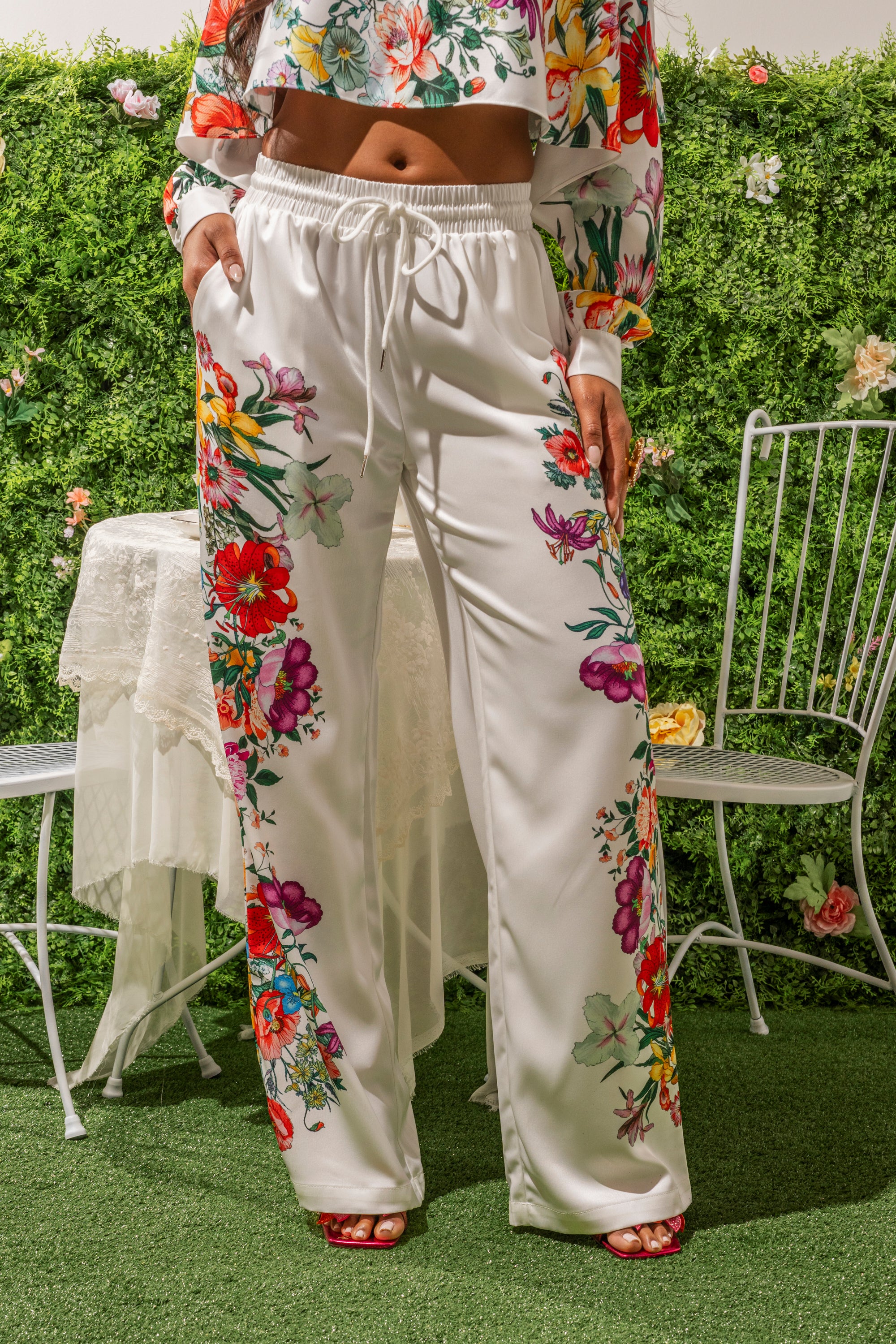 Outdoors on grass, a person wears FULL BLOOM STRAIGHT LEG TROUSERS with colorful floral prints and a matching crop top—a chic spring brunch look. A lace-covered white table and metal chair are surrounded by green foliage and flowers. 1