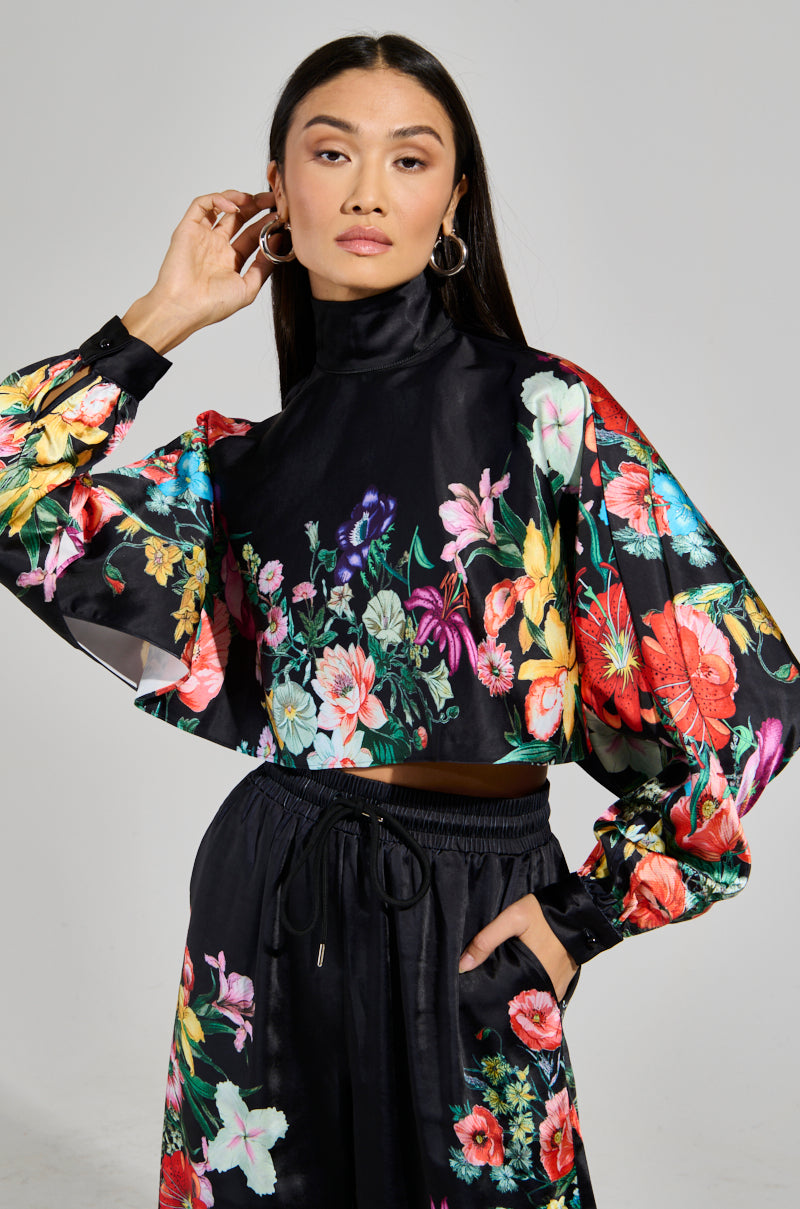 A person confidently poses in the AKIRA Label FULL BLOOM SATIN MOCK NECK BLOUSE, a black satin top adorned with vibrant multicolored floral patterns. They wear matching pants, have long hair, and accessorize with hoop earrings.