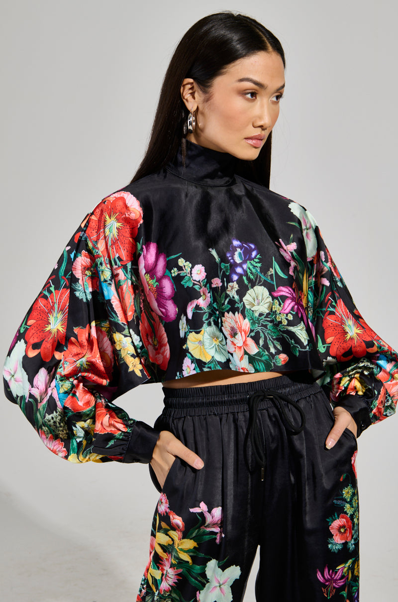 A woman poses confidently in a vibrant floral outfit, wearing the FULL BLOOM SATIN MOCK NECK BLOUSE from the AKIRA Label, adorned with colorful flowers. Her long, dark hair is parted in the middle as she stands with her hands in her pockets against a plain gray background.