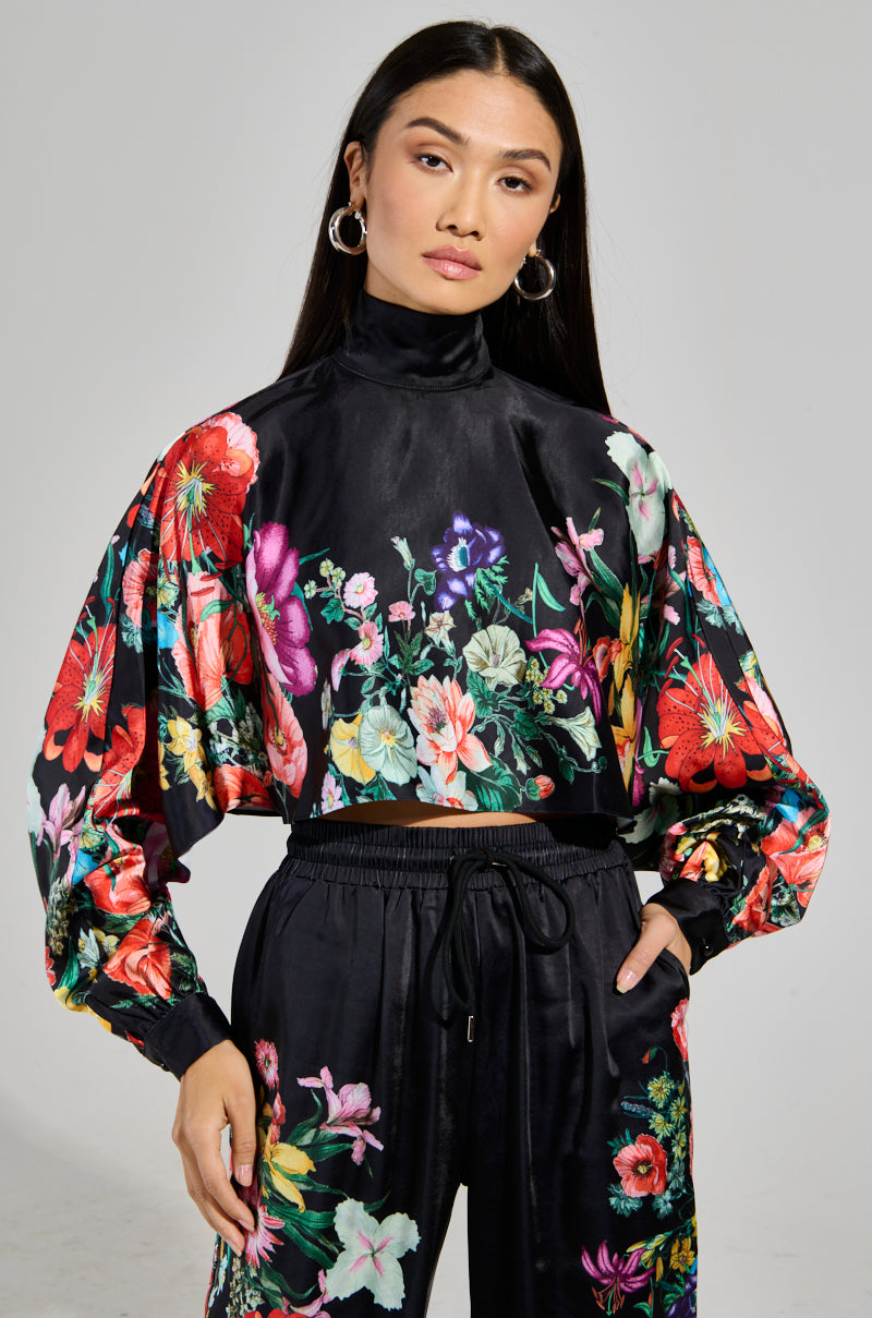 A person with long dark hair dons the FULL BLOOM SATIN MOCK NECK BLOUSE, a black satin piece beautifully adorned with vibrant floral patterns. They accessorize with hoop earrings and pose confidently against a neutral background, highlighting the elegance of this AKIRA Label top.
