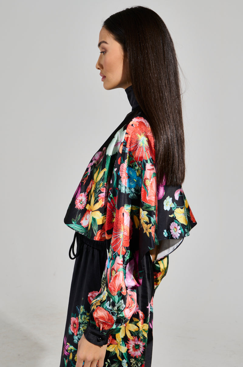 A woman with long dark hair stands in profile, wearing a black dress topped with the Full Bloom Satin Mock Neck Blouse from the AKIRA Label, showcasing floral patterns in red, pink, and green. She is set against a plain light gray background, emphasizing the colorful design of her outfit.