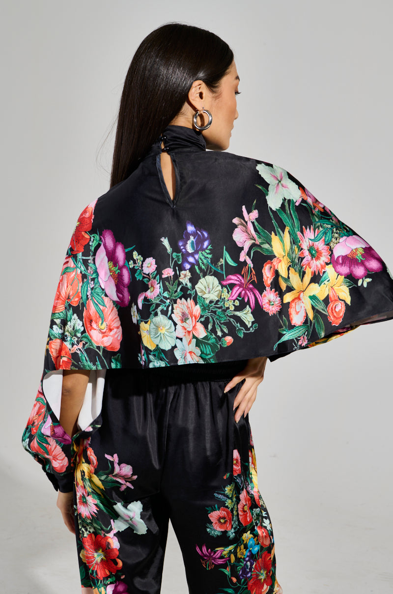 A person with long dark hair is wearing a stylish black outfit, highlighted by the FULL BLOOM SATIN MOCK NECK BLOUSE. This exquisite blouse is adorned with vibrant floral patterns and paired with a flowing cape featuring colorful flowers. They stand against a plain background while gazing to the side.