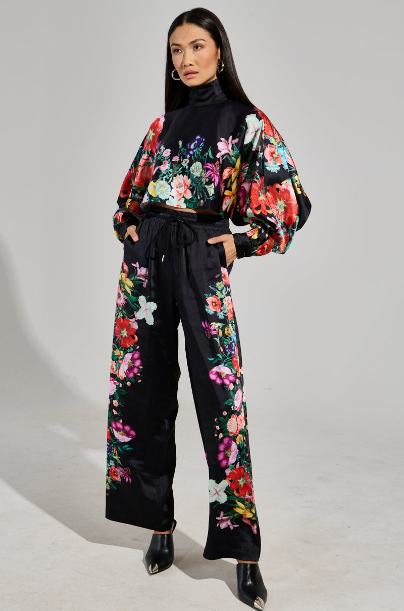 A woman is dressed in an exquisite AKIRA Label ensemble featuring the FULL BLOOM SATIN MOCK NECK BLOUSE along with wide-legged pants, showcasing a striking floral pattern in vibrant red, pink, and green on a black background. With her hands in her pockets, she completes this sophisticated look with stylish black heeled shoes.