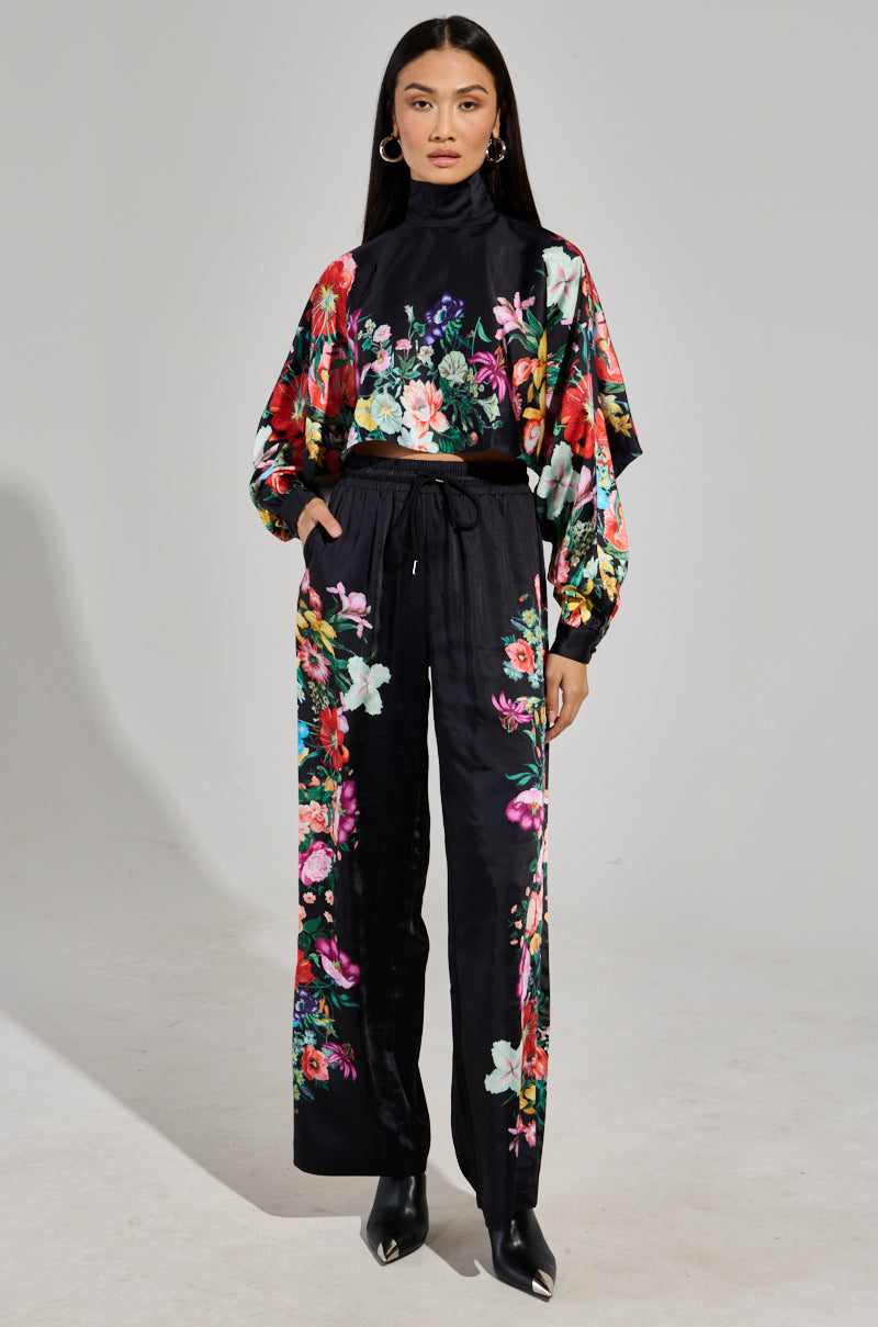 A woman in a floral outfit stands confidently, sporting the AKIRA Label FULL BLOOM SATIN MOCK NECK BLOUSE featuring a vibrant floral pattern on a black background. Her hands rest casually in her pant pockets, paired with hoop earrings and black shoes, completing the chic ensemble.