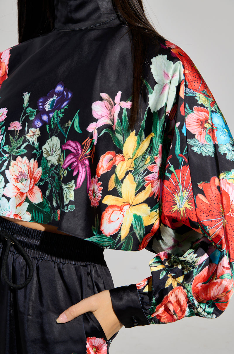 A person wearing the FULL BLOOM SATIN MOCK NECK BLOUSE, which features vibrant flowers such as lilies and daisies on a sleek black background, truly stands out.
