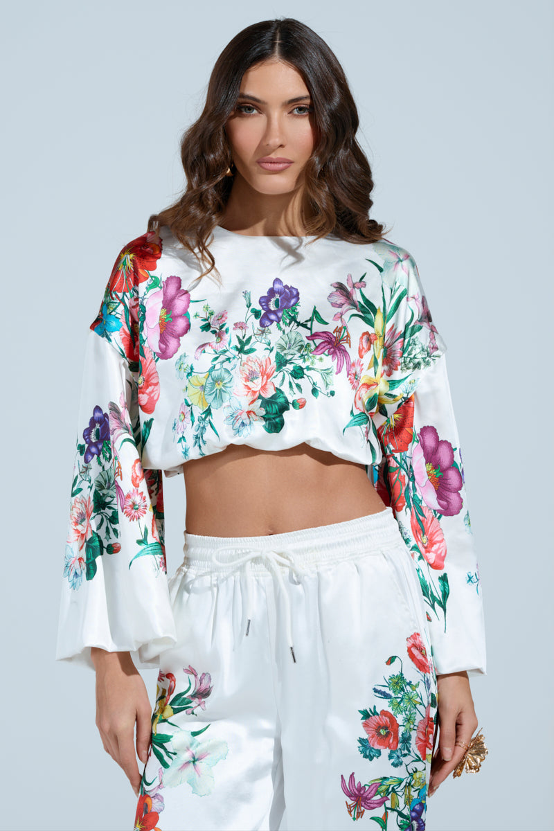 A woman with long, wavy hair wears the FULL BLOOM ZIP OFF SLEEVES BLOUSE and matching pants, featuring a floral print. The vibrant flowers and convertible puff sleeves stand out against the plain light blue background.