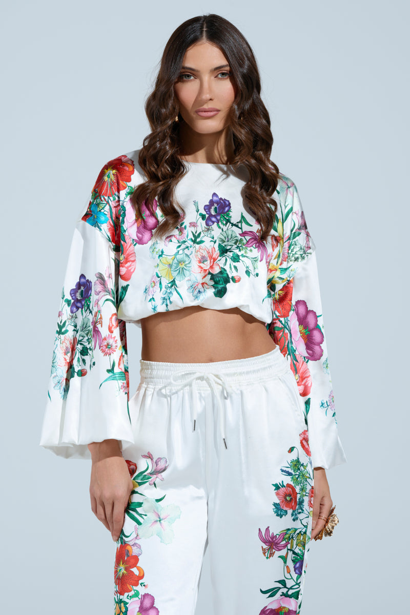 A woman with long wavy hair wears a satin floral-patterned outfit. The FULL BLOOM ZIP OFF SLEEVES BLOUSE features wide, convertible sleeves with vibrant flowers, while her matching pants have floral designs near the pockets on a light blue background.