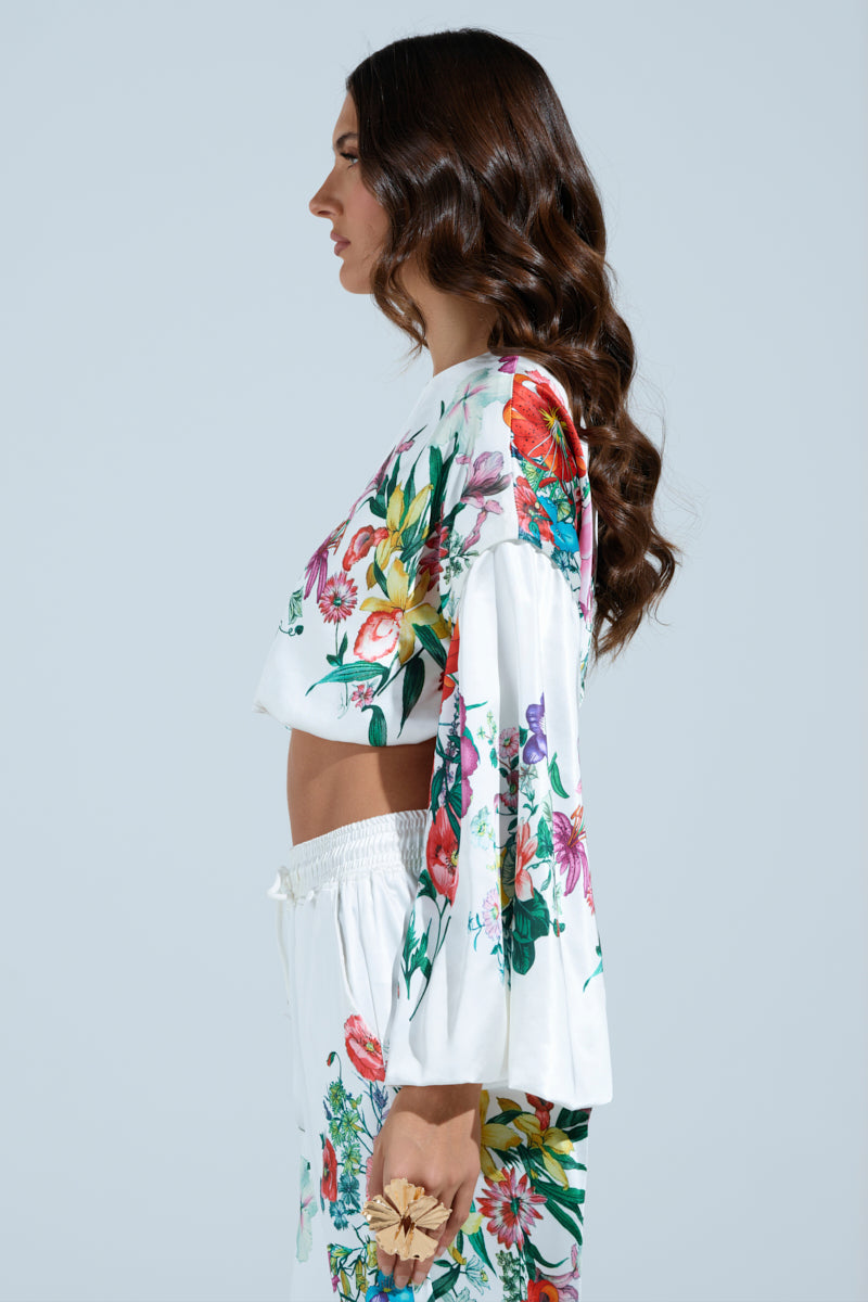 A woman with long brown wavy hair stands in profile against a light background. She wears the FULL BLOOM ZIP OFF SLEEVES Blouse, a floral print satin crop blouse and pants with convertible puff sleeves. In one hand, she holds a delicate flower accessory.