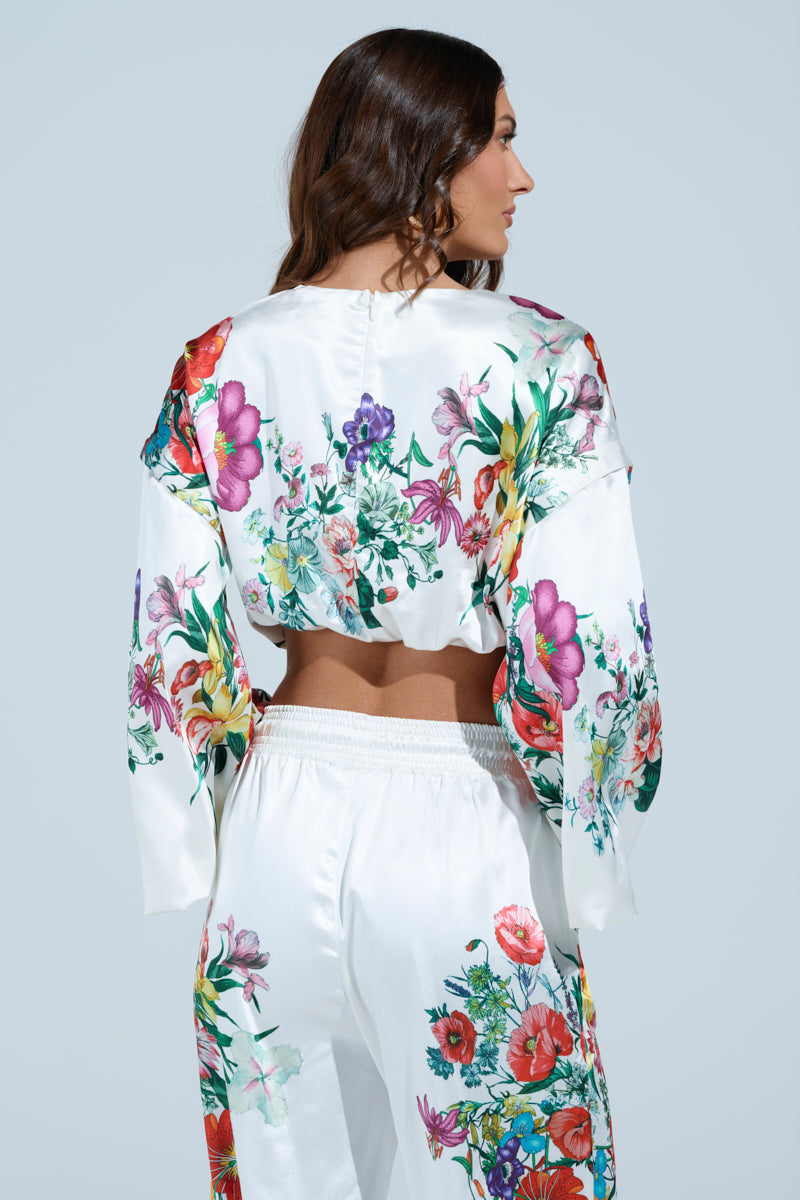 A person with long hair wears the chic FULL BLOOM ZIP OFF SLEEVES BLOUSE, a white floral print satin crop top with convertible puff sleeves. Colorful flowers pop as they turn left, showcasing the blouse against a light blue background.
