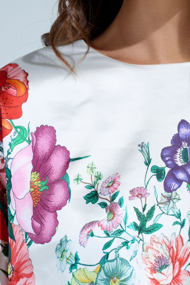 A close-up shows a person wearing the FULL BLOOM ZIP OFF SLEEVES BLOUSE, featuring vibrant pink, purple, red, and orange floral prints with green leaves on a white satin crop. Convertible puff sleeves frame their wavy brown hair.