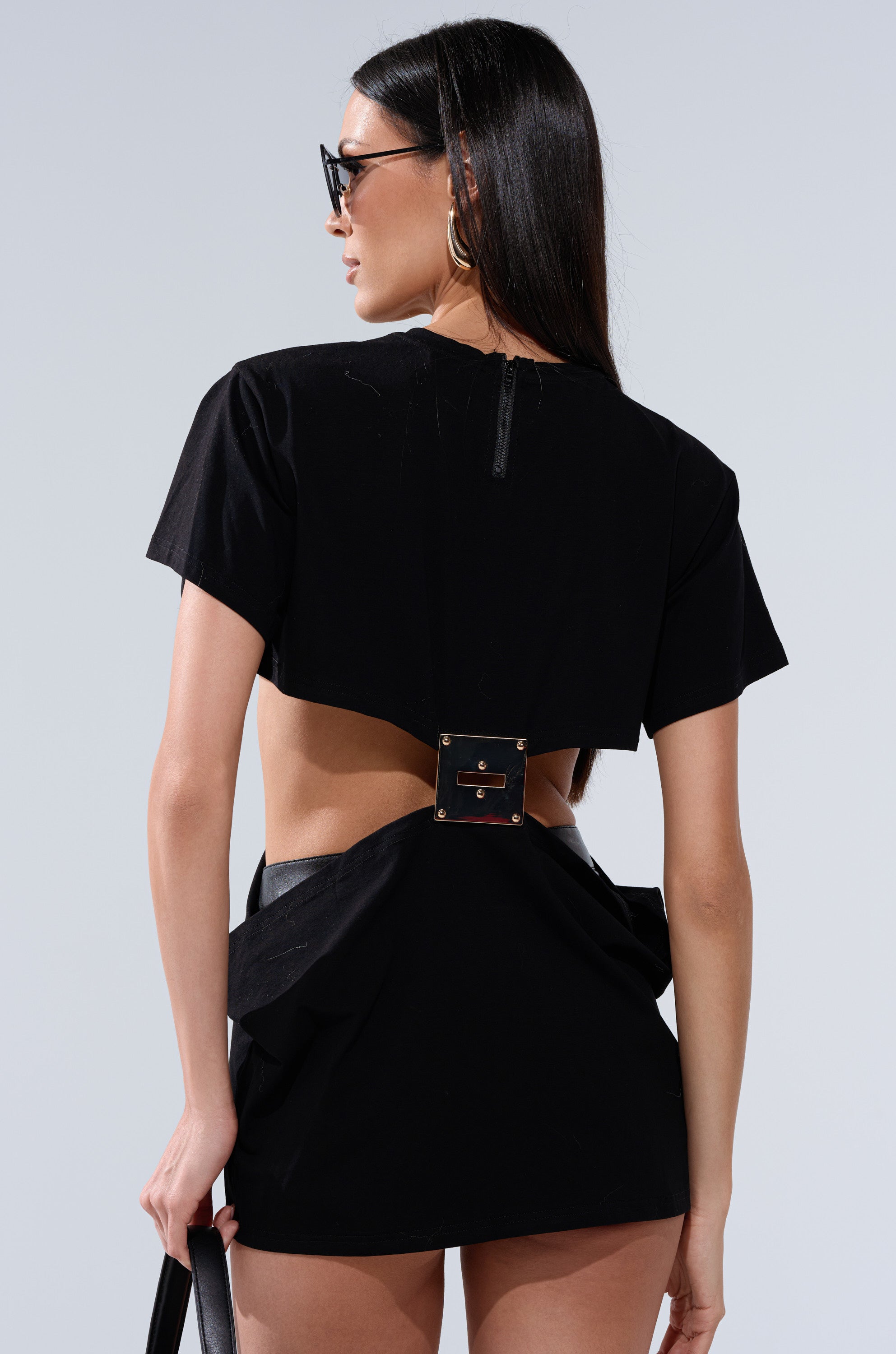 A woman with long dark hair wearing sunglasses and the I'M THE BOSS CUT OUT TSHIRT, featuring an open back, metallic buckle detail, and cut-out design, holds a black handbag while facing away from the camera.