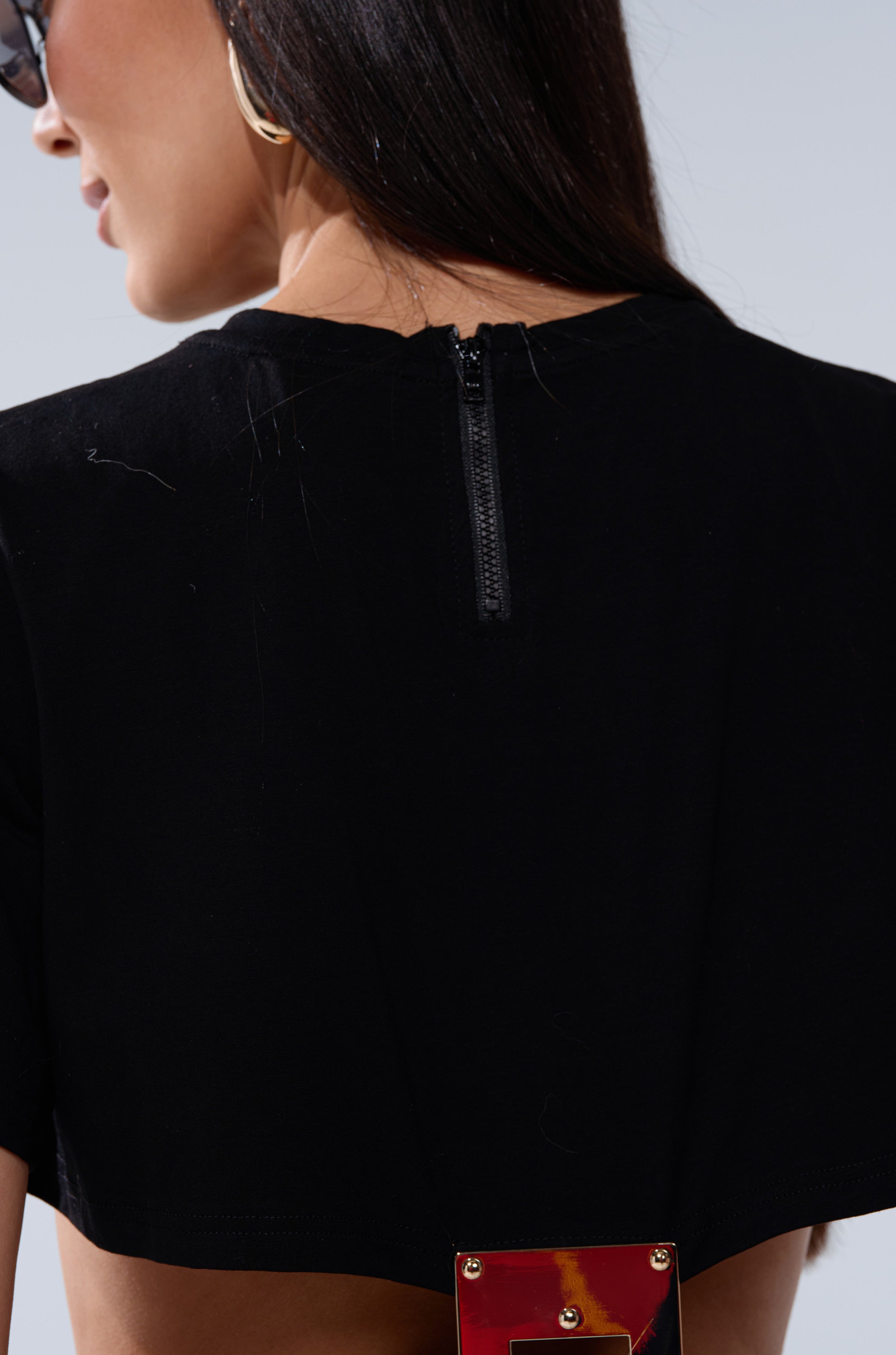 A woman with long dark hair wears sunglasses and gold hoop earrings, shown from behind in the AKIRA Label I'M THE BOSS CUT OUT TSHIRT featuring a visible back zipper and metal clasp detail at the bottom.