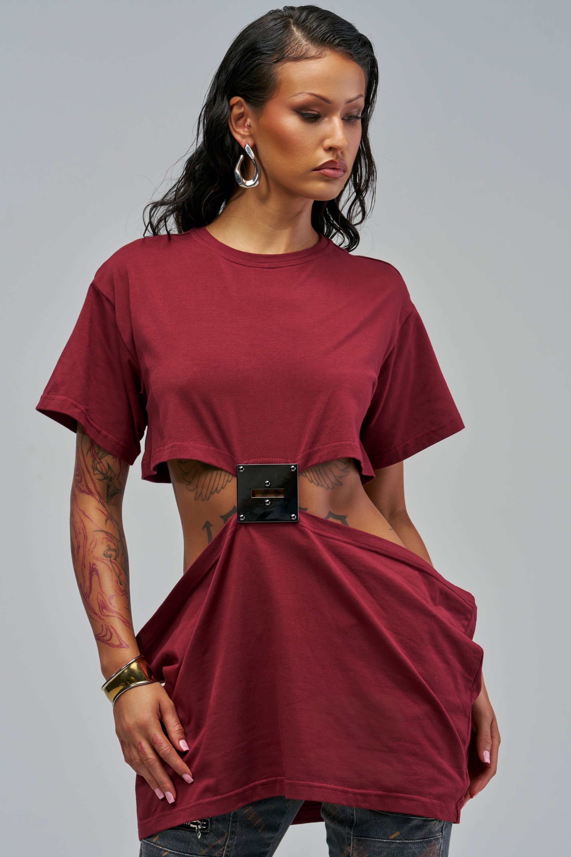 A person wearing a burgundy BUCKLE UP BABE CUTOUT TEE with a cropped short-sleeve silhouette and a central rectangular buckle detail at the waist.