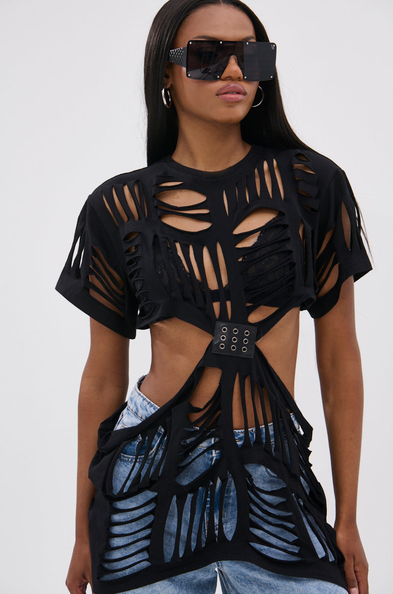 A woman in grunge fashion wears oversized black sunglasses, hoop earrings, and the SHREDDED OVERSIZED SHORT SLEEVE TEE with a grommet belt detail, paired with blue jeans, standing against a plain light background.