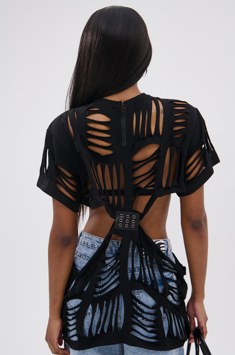A person with long, straight hair wears the SHREDDED OVERSIZED SHORT SLEEVE TEE, featuring black fabric with unique back and sleeve cutouts, paired with blue jeans. The rear view showcases this grunge-inspired style’s standout pattern.